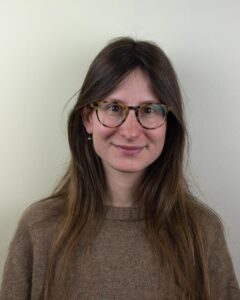 A portrait of a person with long brown hair and tortoiseshell eyeglasses, smiling slightly while looking at the camera. They are wearing a brown knit sweater and standing in front of a light-colored wall. The lighting is soft and even, giving the image a warm and approachable feel.