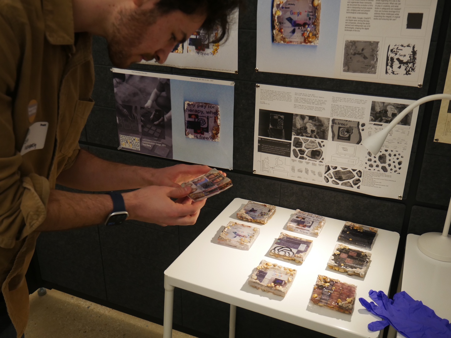 A person in a tan shirt examines a small resin-cast tile containing mixed-media imagery, part of a larger display on a white table. Nine handmade tiles are laid out in rows, each embedding digital fragments, text, and electronics into translucent surfaces. Behind the table, printed posters feature scanned designs, research notes, and magnified images. A bright desk lamp and blue nitrile gloves are also visible, suggesting an interactive or delicate handling environment.