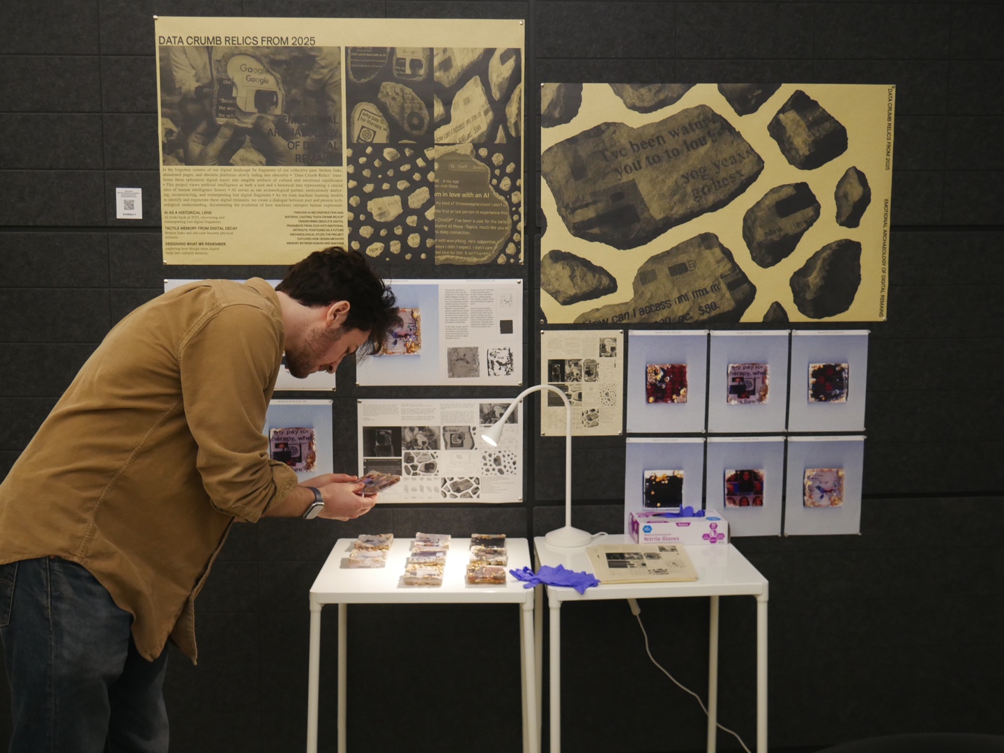 Close-up view of a white table displaying nine resin-cast data relic tiles embedded with text, circuitry, and visual fragments. A desk lamp illuminates the arrangement, while a box of nitrile gloves, a pair of blue gloves, and a vintage magazine are placed on the adjacent table. Posters pinned on the wall explain the project and show reference materials, including digital decay imagery and speculative designs.
