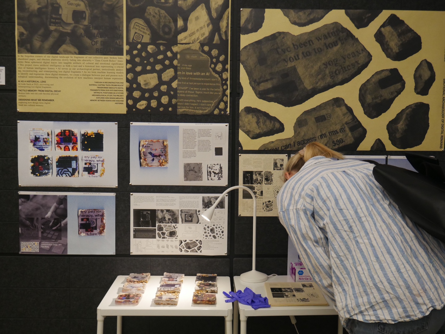 Wider view of the exhibit installation titled “Data Crumb Relics from 2025.” A person in a tan shirt examines one of the resin-cast tiles on a lit table. The wall behind features enlarged posters with gold-toned graphics of digital artifacts preserved as speculative archeology. Titles on the posters read “DATA CRUMB RELICS FROM 2025” and “DESIGNING WHAT WE REMEMBER.” The space blends research, critical media, and art-based inquiry into digital preservation.