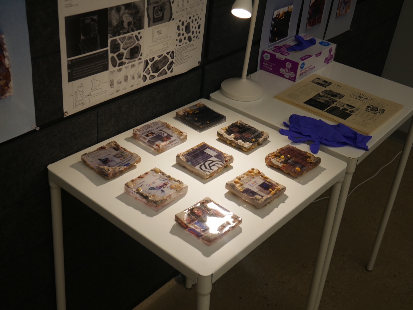 A visitor in a blue-and-white striped shirt leans over to closely examine a series of resin-cast digital relic tiles on a table. Behind them, posters titled “Data Crumb Relics from 2025” showcase conceptual renderings of digital decay and memory, with visual metaphors of technological erosion. A desk lamp lights the table, which also holds gloves and printed ephemera. The installation is designed to evoke tactile interaction and speculative history.