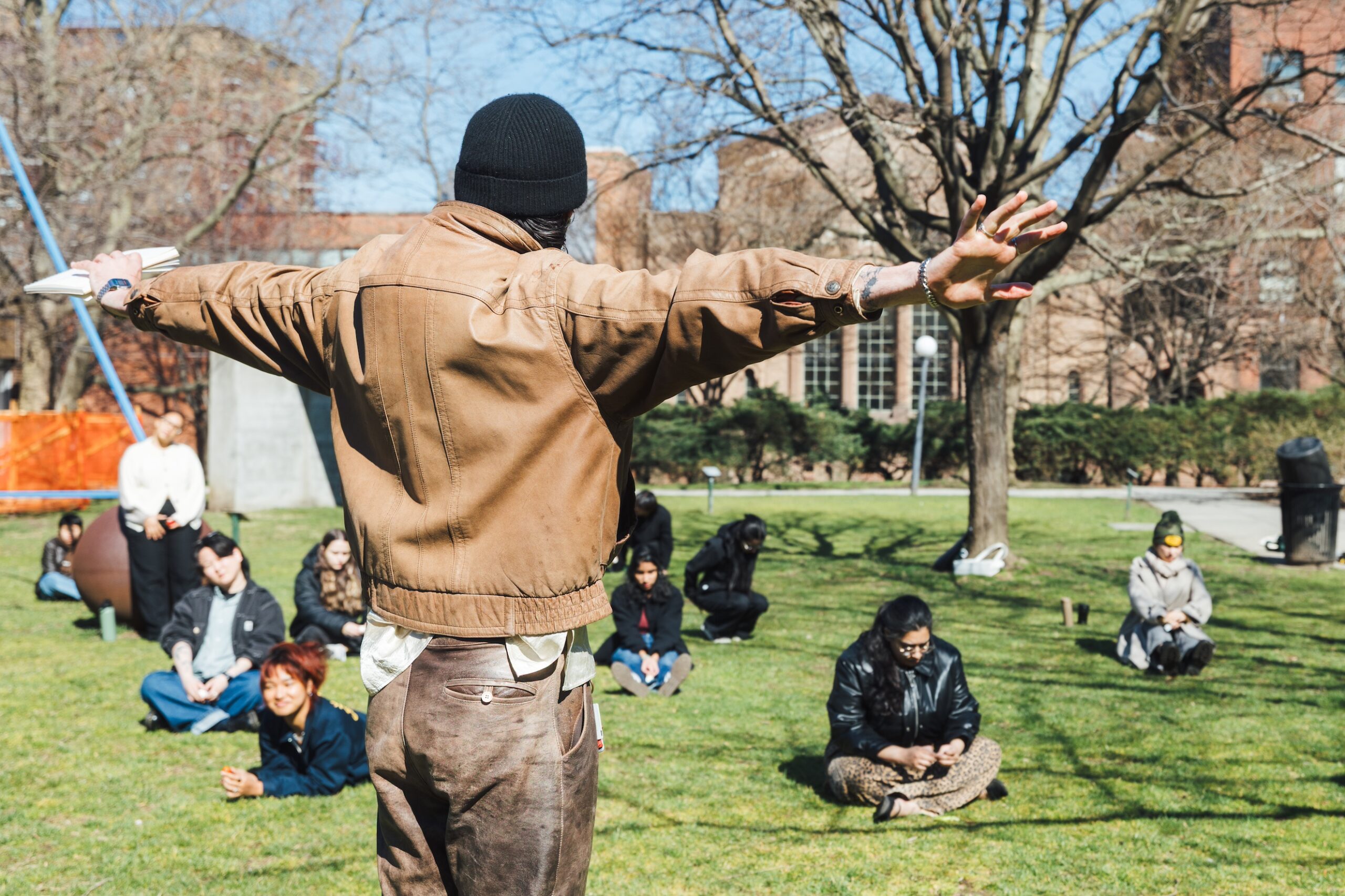 Students sit and lay on a campus lawn in the sun behind a facilitator, their back to the camera and arms outstretched to their sides