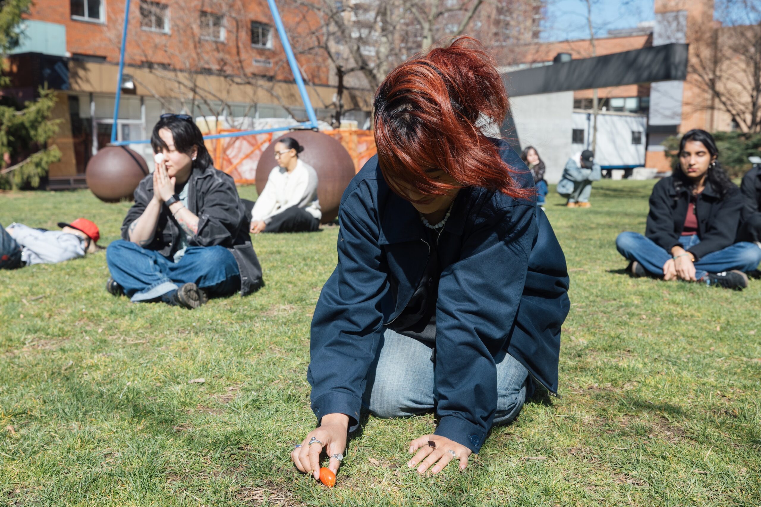 A student kneeling and smiling to themselves on the grass while they hold a cherry tomato, with students behind them, eyes closed in different sitting positions