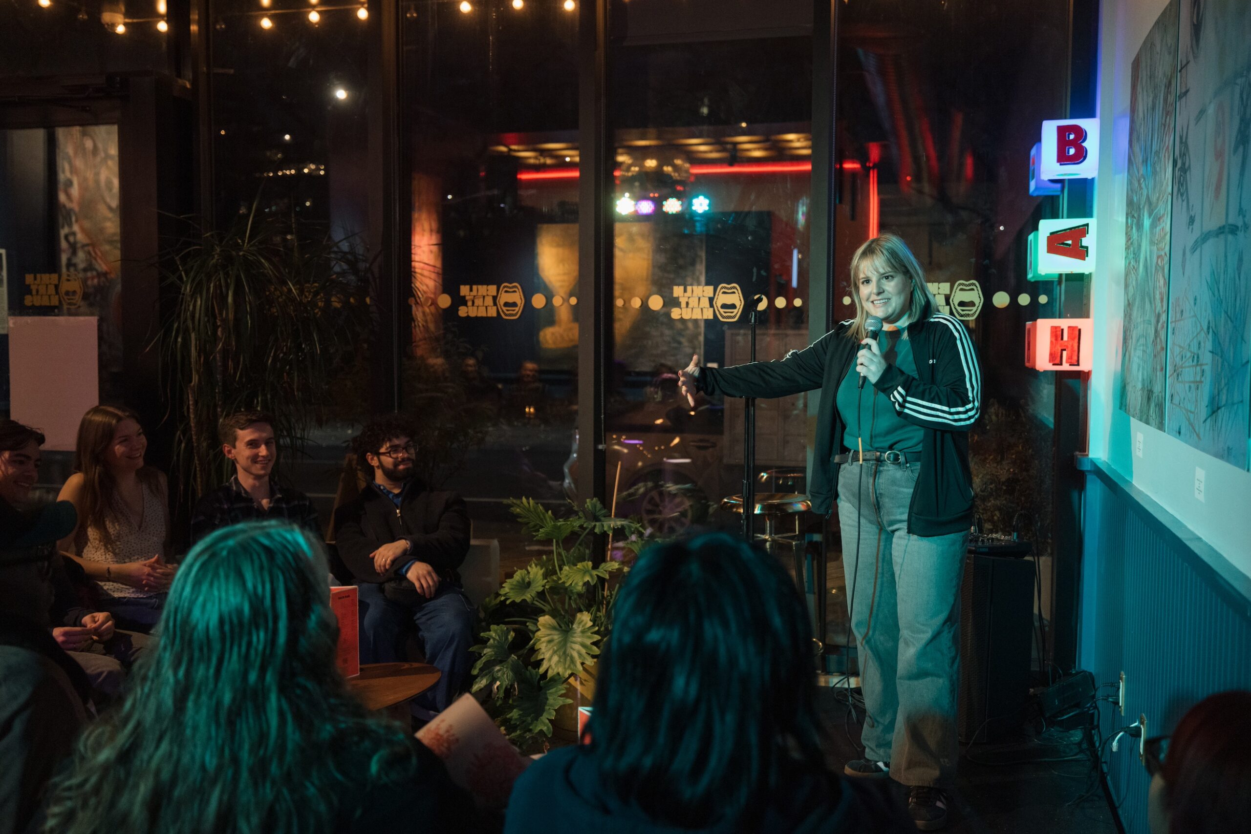 Kath Barbadoro on stage holding a microphone during a stand-up comedy act, gesturing at the audience