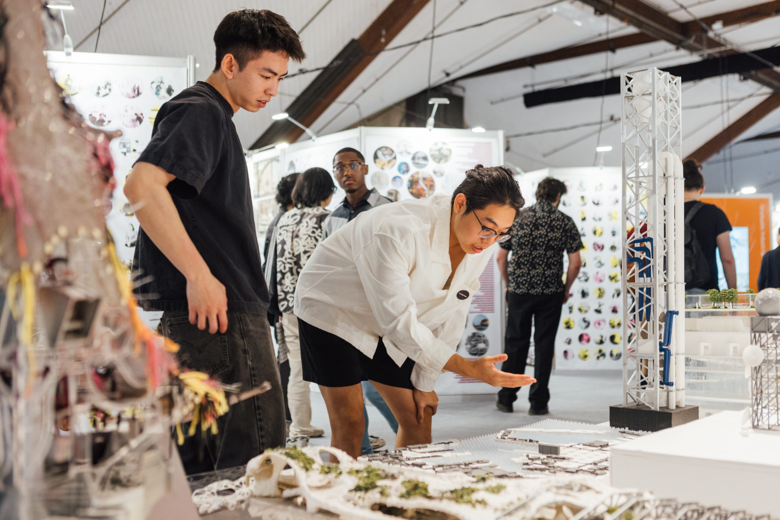 Two people at the Design Show are looking closely at someone's work. All around them are other exhibitions of work by students.
