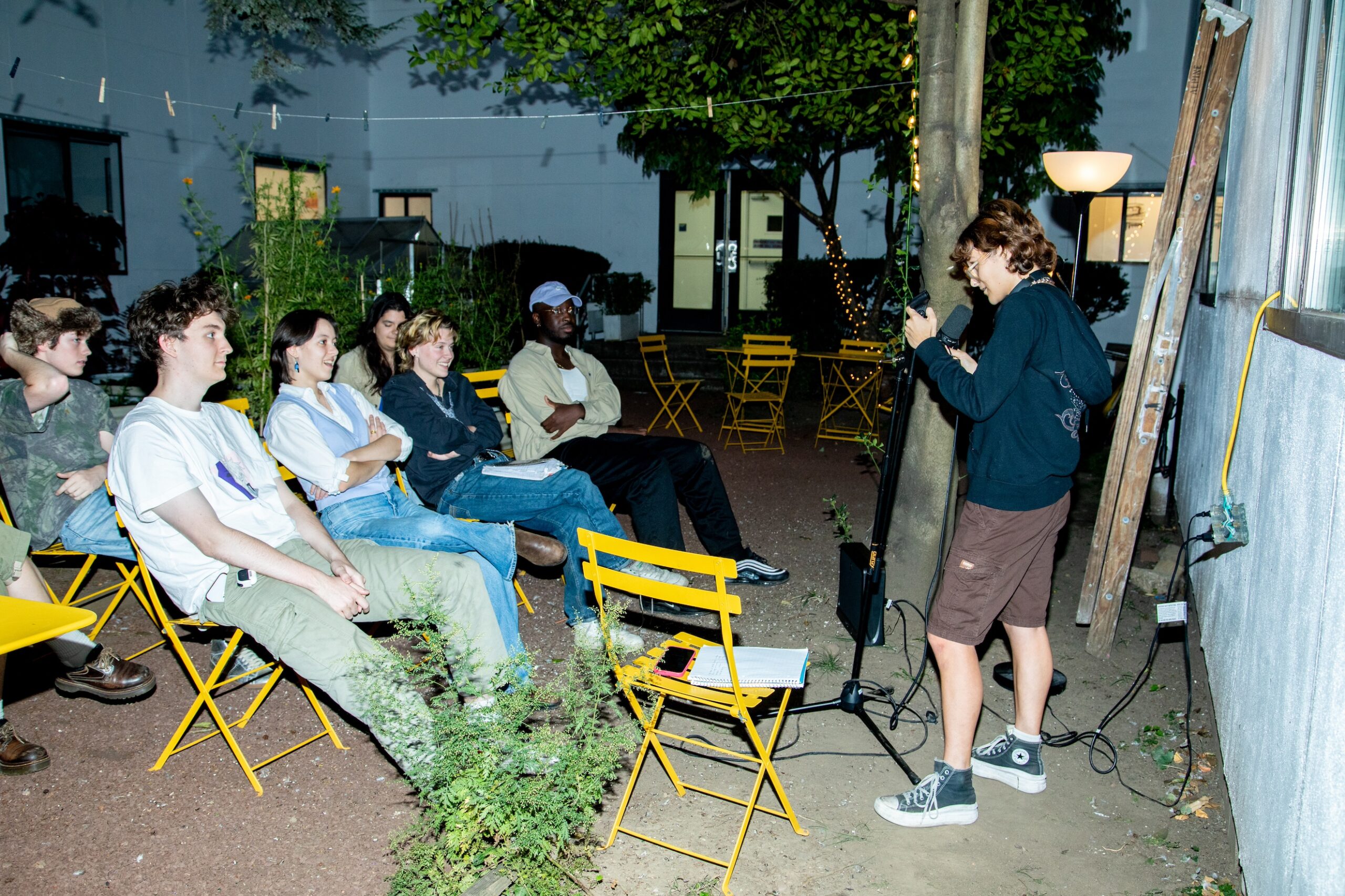 A small audience of students sits in a courtyard, watching a stand-up comedy act by one of their peers