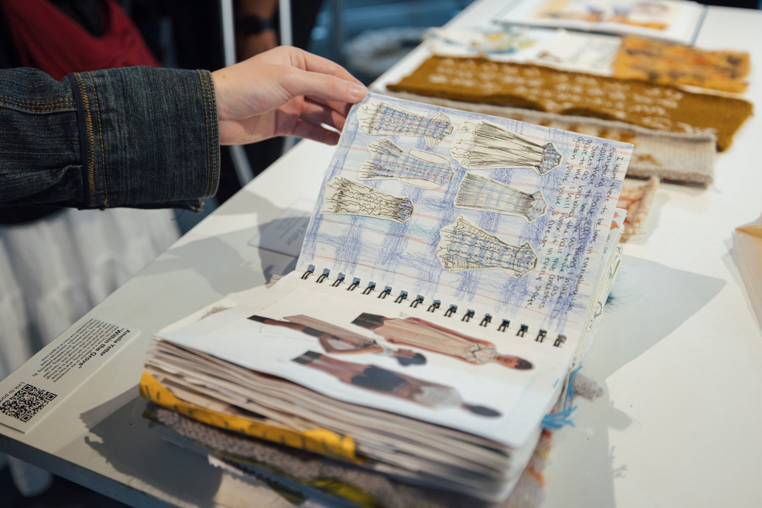 A close-up view of a spiral-bound fashion design sketchbook held open on a white table. A person wearing a denim jacket gently lifts the top page, which features detailed dress sketches over a blue and red grid background, accompanied by handwritten notes. The dresses vary in silhouette and feature elements like pleats, smocking, and texture variations. The page underneath shows cut-out illustrations of runway-style fashion figures. In the background, textile samples in shades of brown, gold, and cream are laid out on the table, partially out of focus.