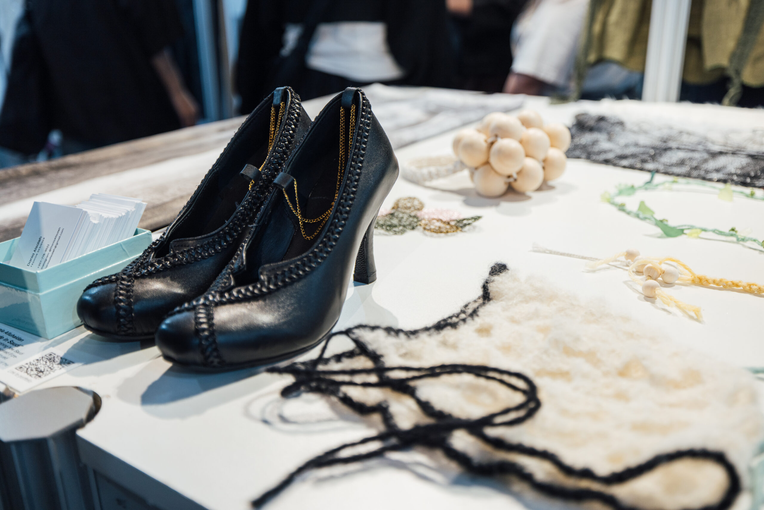 A pair of black high-heeled shoes with braided leather trim and delicate gold chain accents on display atop a white table. To the left, a teal box holds a stack of printed business cards. Scattered across the table are various small design samples and textile experiments, including a bunch of smooth wooden beads, swatches of black lace and white wool, braided cords, and embroidered floral motifs. Blurred figures in dark clothing and exhibition panels are visible in the background, suggesting the setting is a student or designer showcase.