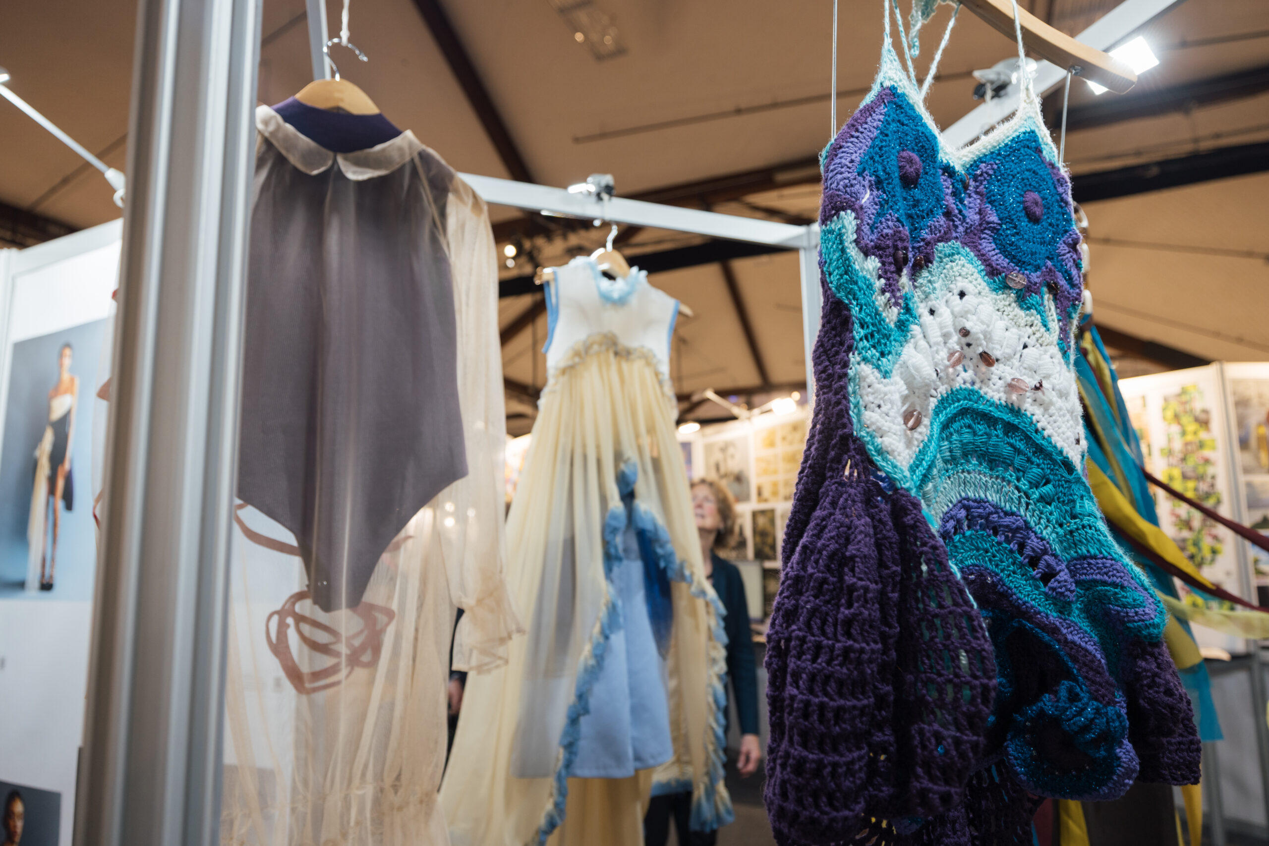 A trio of avant-garde garments hang on wooden hangers from a metal display structure at a fashion exhibition. In the foreground on the right, a vibrant crochet dress features bold, textured patterns in purple, teal, white, and blue yarns, with floral-like motifs and open-stitch details. To the left, a translucent beige bodysuit with a pointed hem and sheer long sleeves is layered over a darker fabric and finished with a delicate collar. At the center, a sleeveless dress made of sheer golden-yellow fabric is adorned with soft blue embellishments and a sculptural blue ruffle cascading down the front. Exhibition boards, lighting, and attendees are visible in the softly blurred background.