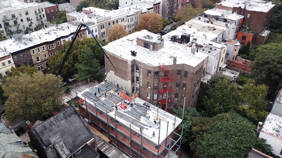 Aerial photo of a mid-construction building site in a Brooklyn residential neighborhood. The image shows the steel framework of the new structure adjacent to an existing brown brick apartment building with red fire escapes. A crane is in operation, and construction workers are visible on the site. Surrounding the site are rows of older buildings and dense tree canopies.