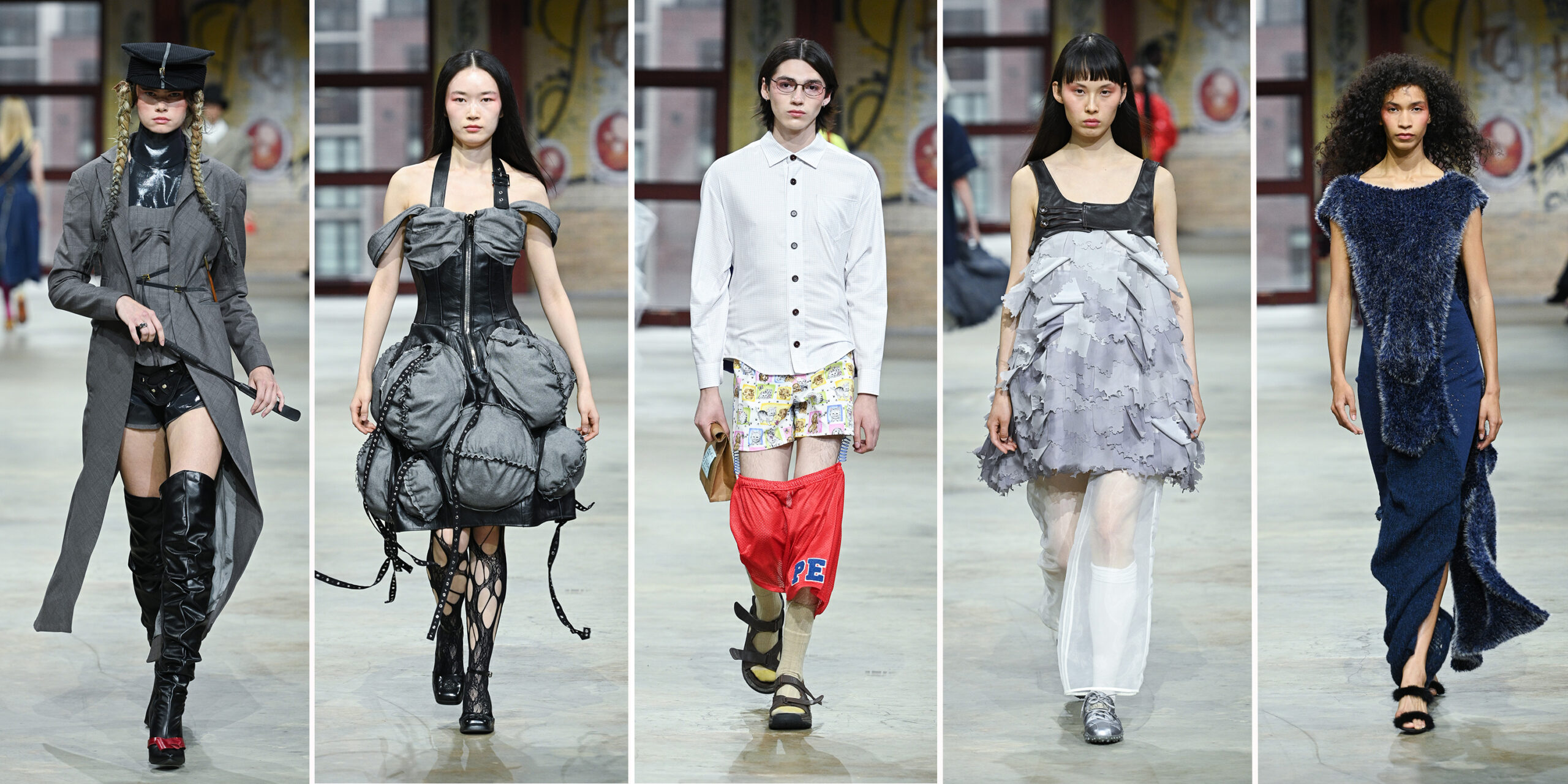 A lineup of five models walking the runway in an eclectic, experimental fashion collection, set against a graffiti-covered brick backdrop. From left to right: The first model wears a structured grey military-style coat with black buttons and straps, layered over glossy black latex and thigh-high black patent leather boots with a red bow accent. A black military cap and braided pigtails complete the look. The second model wears a sculptural, charcoal grey strapless dress with dramatic ballooned ruching, a leather bodice, and black lace-up cutout boots. The third model wears a crisp, button-down white shirt paired with playful, cartoon-print boxer shorts and red mesh athletic shorts slouched at the knees. Beige socks, sport sandals, and a paper-wrapped book add to the conceptual styling. The fourth model wears a sleeveless grey mini dress with cascading, petal-like layers and a black leather yoke. Sheer white pants peek out beneath the hem, paired with metallic silver sneakers. The fifth model wears a deep navy blue knit dress with asymmetrical, fuzzy-textured panels that wrap around the body. The dress features a side slit and is styled with black fuzzy heels.