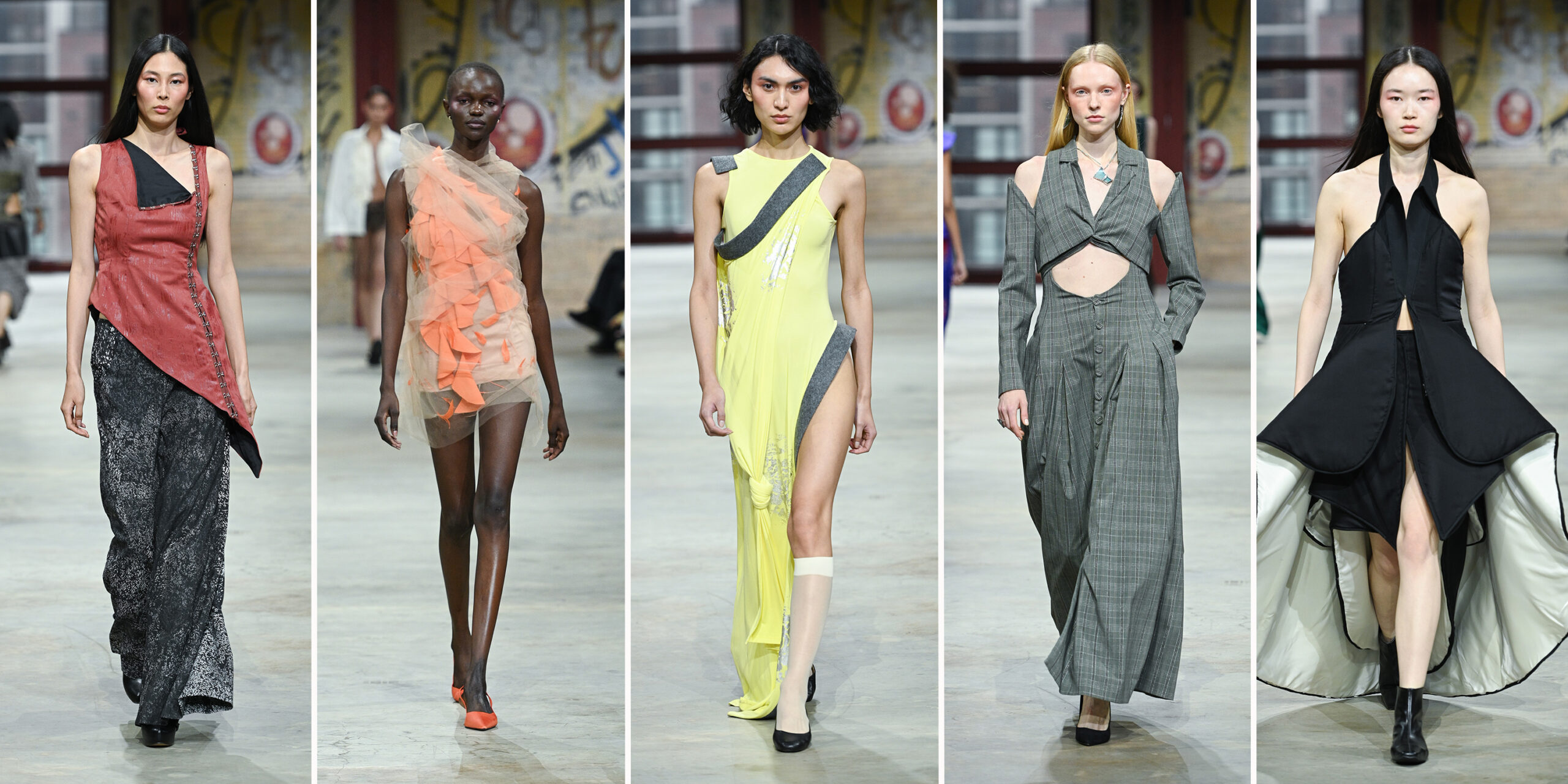 Five models walk a fashion show runway, each wearing avant-garde garments in muted and vibrant tones, with a graffiti-covered wall in the background. From left to right: The first model wears an asymmetrical red and black sleeveless tunic with side button detailing, layered over wide-leg black pants featuring a mottled silver pattern. The second model wears a sheer one-shoulder mini dress made from layered peach and tangerine organza, paired with color-blocked red and orange shoes. The third model wears a dramatic sleeveless dress in bright chartreuse with gray trim and a thigh-high slit, detailed with printed elements and gathered knots. The look is styled with white socks and black flats. The fourth model wears a tailored gray plaid ensemble featuring a cut-out top with a collar and a buttoned high-waisted skirt. A blue pendant necklace adds contrast to the sharp structure. The fifth model wears a black sculptural halter-neck dress with a voluminous, architectural silhouette and high slits, revealing white fabric lining and black ankle boots.
