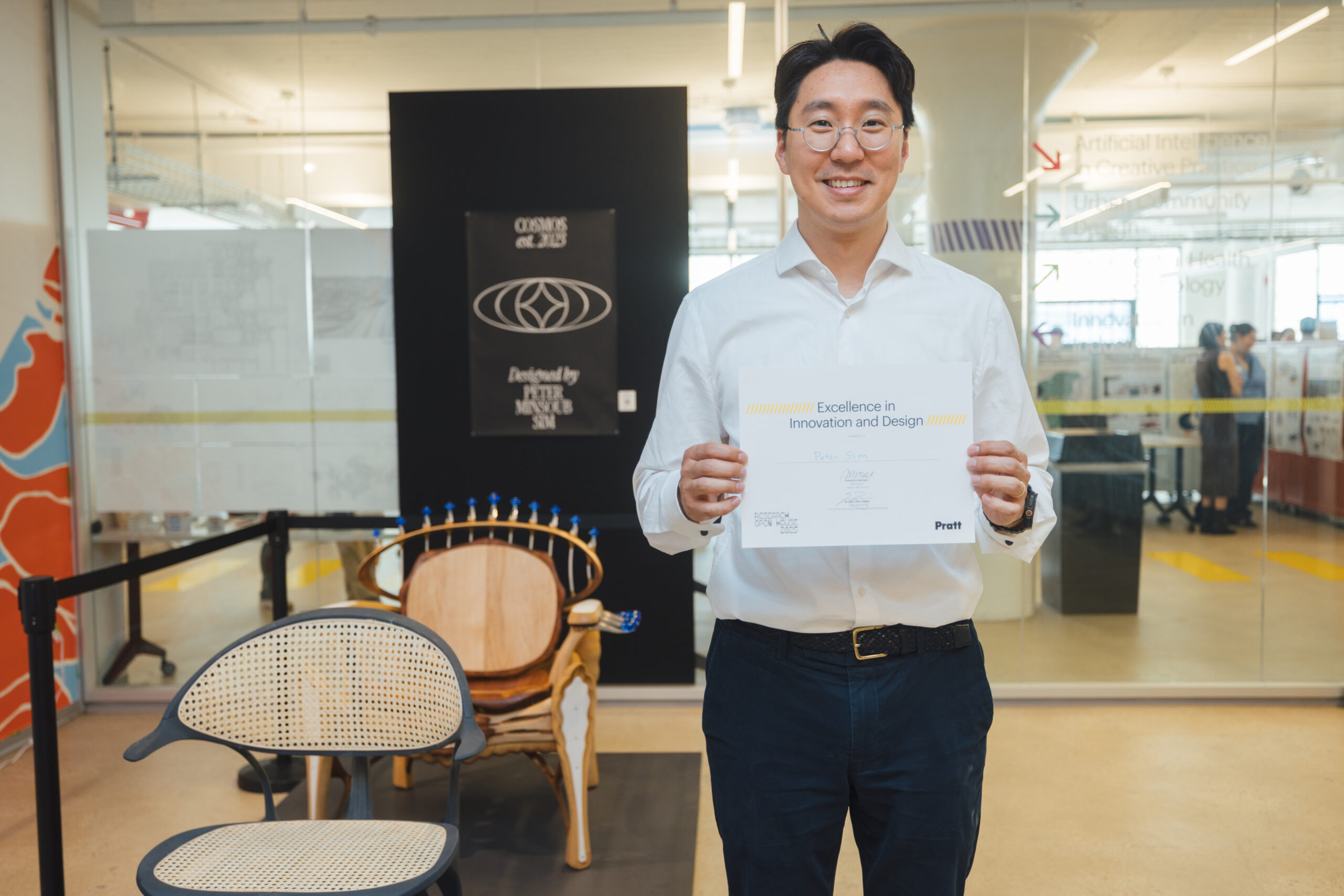 A student smiles while holding a certificate for “Excellence in Innovation and Design” at Pratt Institute’s Research Open House. Behind him is a glass-walled exhibition space and a display of chairs, one with a futuristic design and the other made from repurposed materials.
