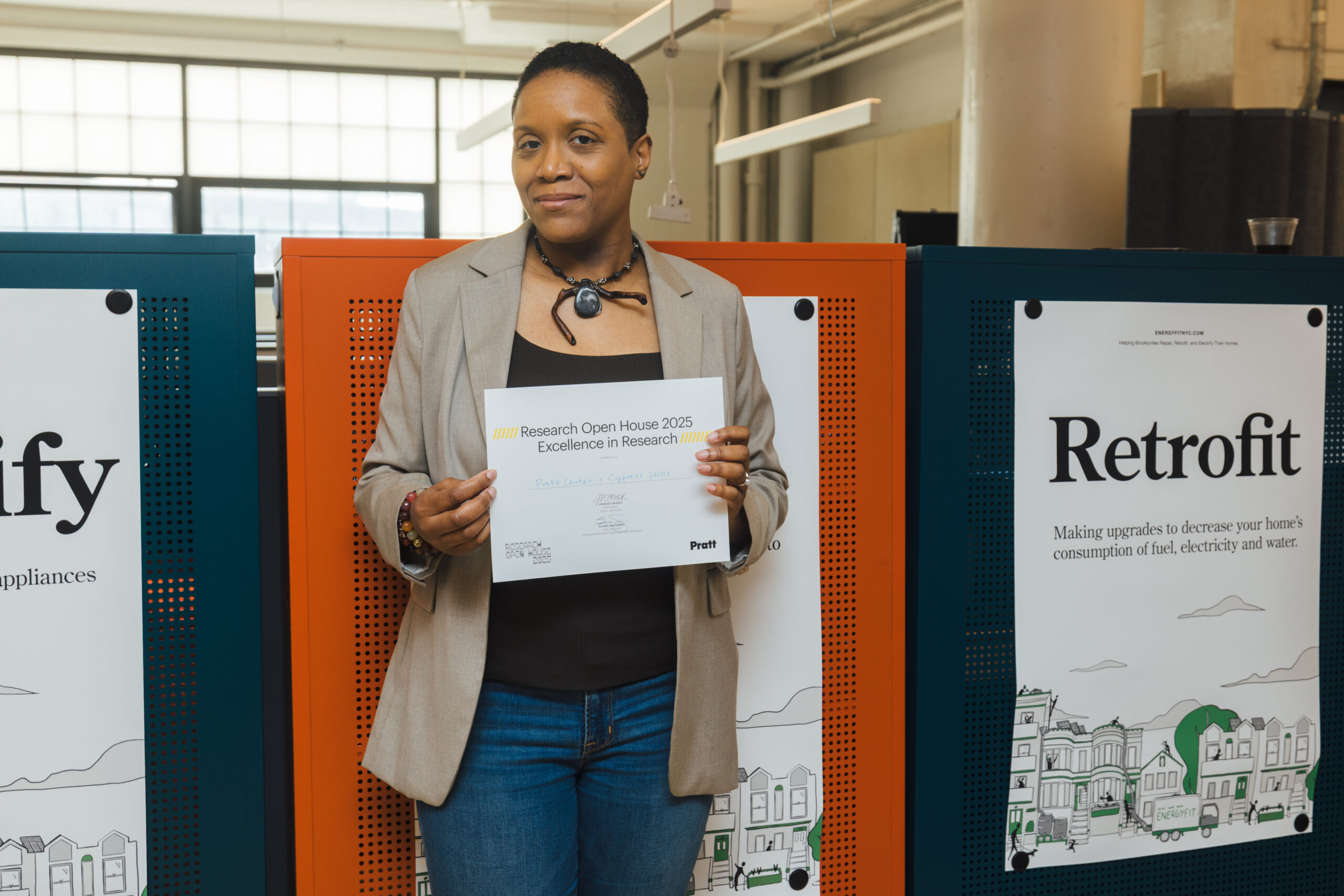 A person holds a certificate for “Excellence in Research” at Pratt Institute’s 2025 Research Open House. They stand in front of a colorful display panel with informational posters, one of which reads “Retrofit,” detailing energy-efficient home upgrades.