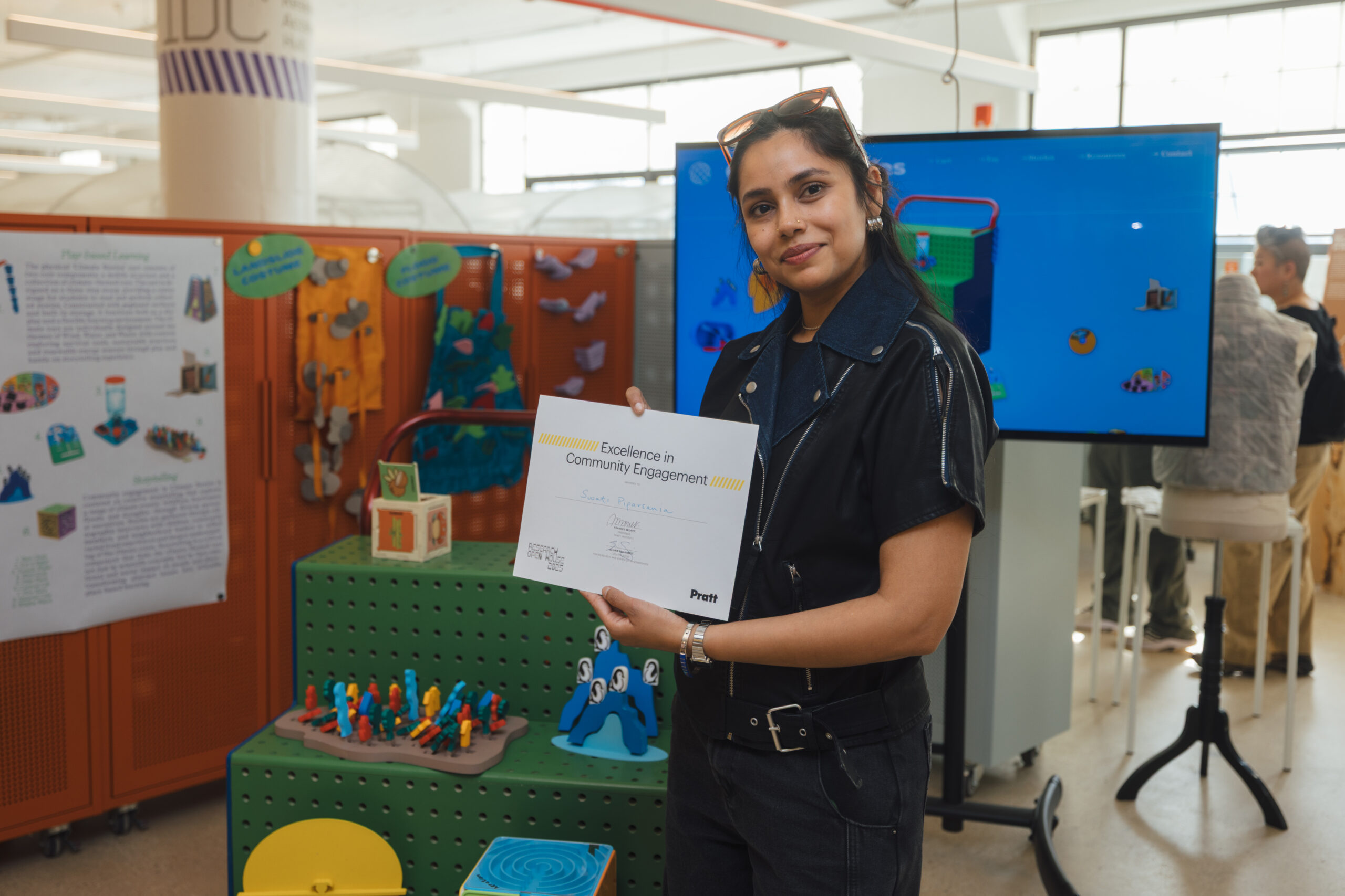 A student stands indoors holding a certificate for “Excellence in Community Engagement” at Pratt Institute’s Research Open House. Behind her is a colorful display of interactive design projects, including 3D models and educational materials.