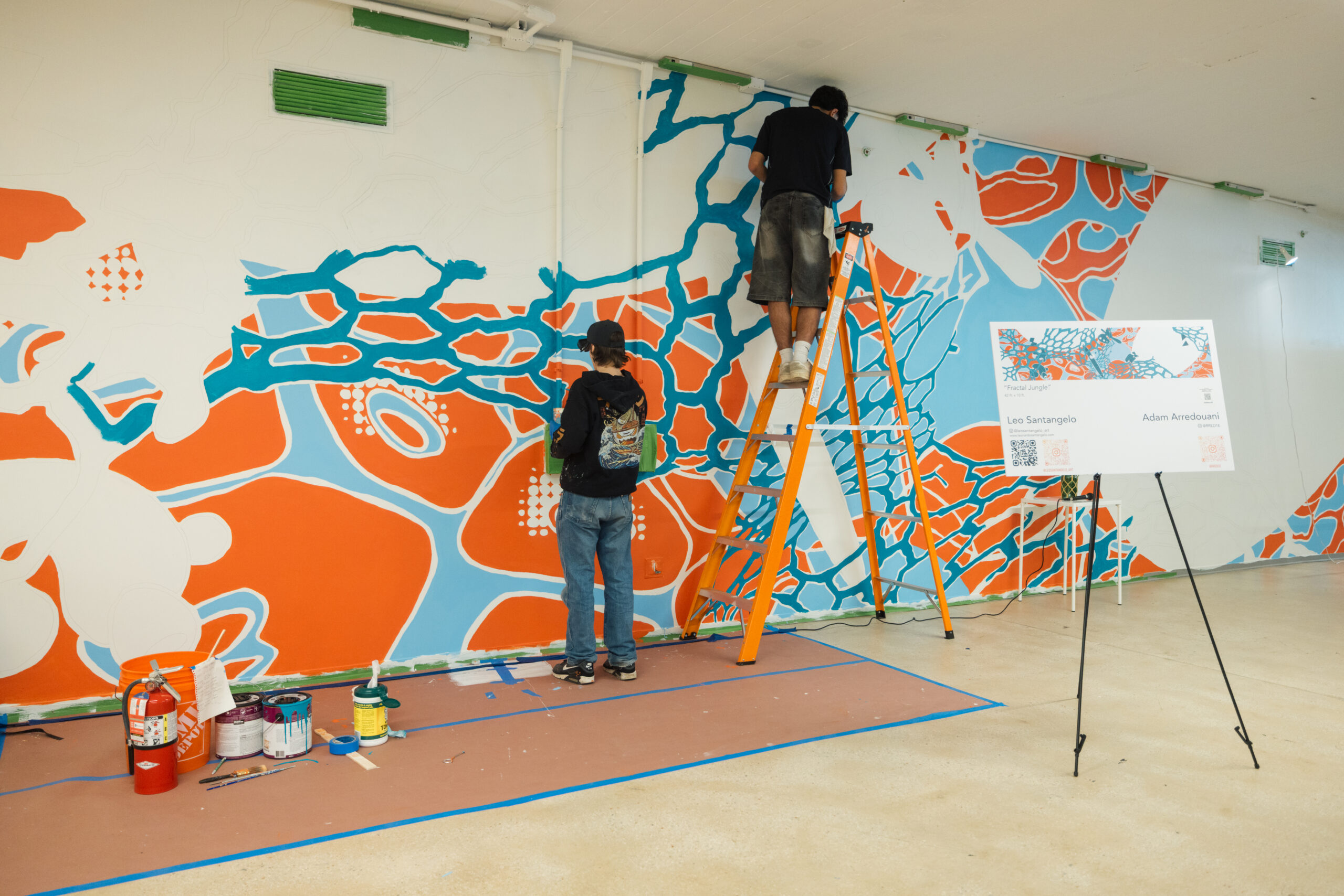 Two artists work on painting a large, vibrant mural featuring bold red, blue, and white abstract organic shapes. One artist stands on a ladder while the other paints at ground level. Paint cans and supplies are scattered on a protective floor covering, and a sign nearby identifies the mural as Fractal Jungle by Leo Santangelo and Adam Arredouani.