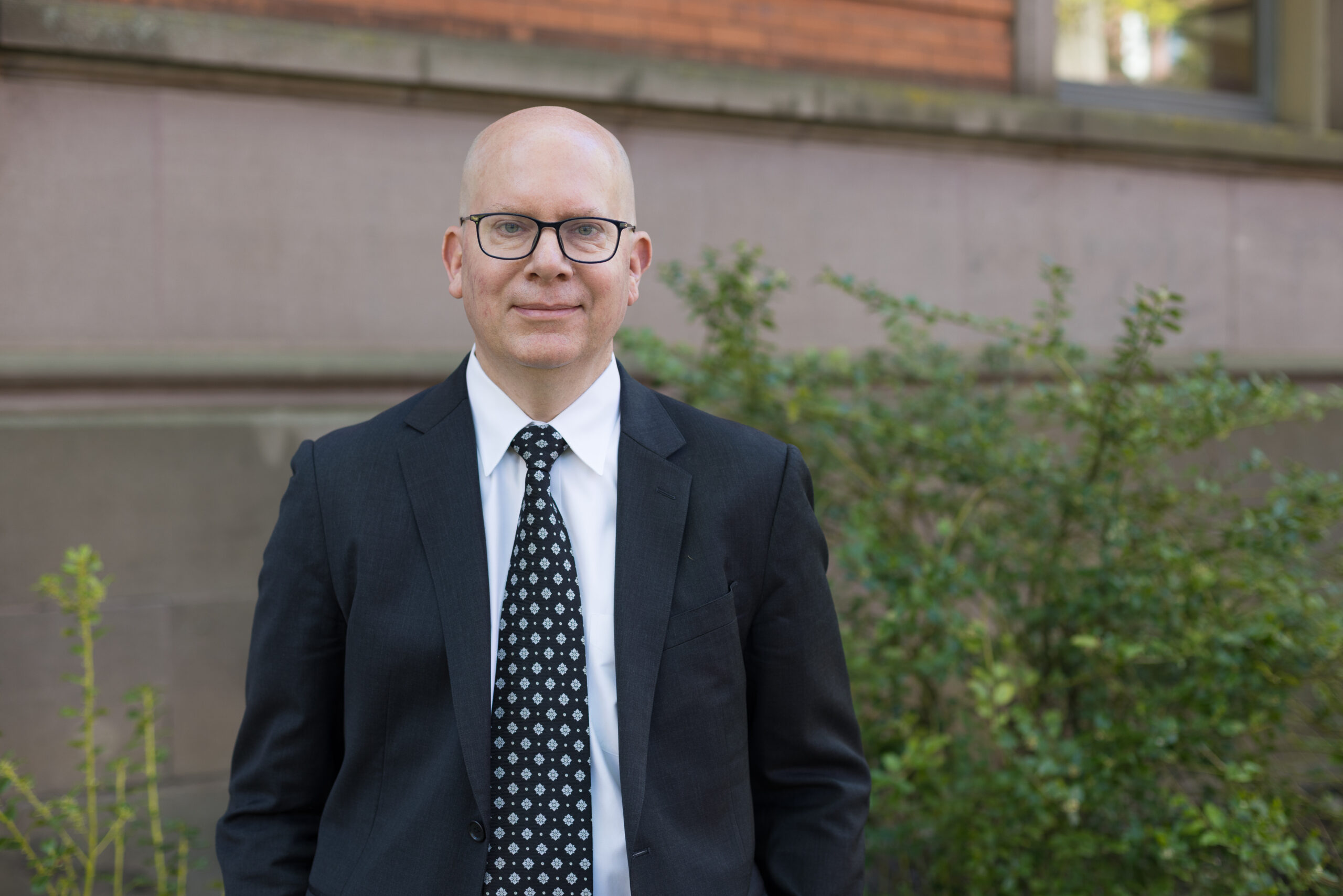 A man stands in front of a building. He wears a suit and tie and glasses. There is greenery behind him.