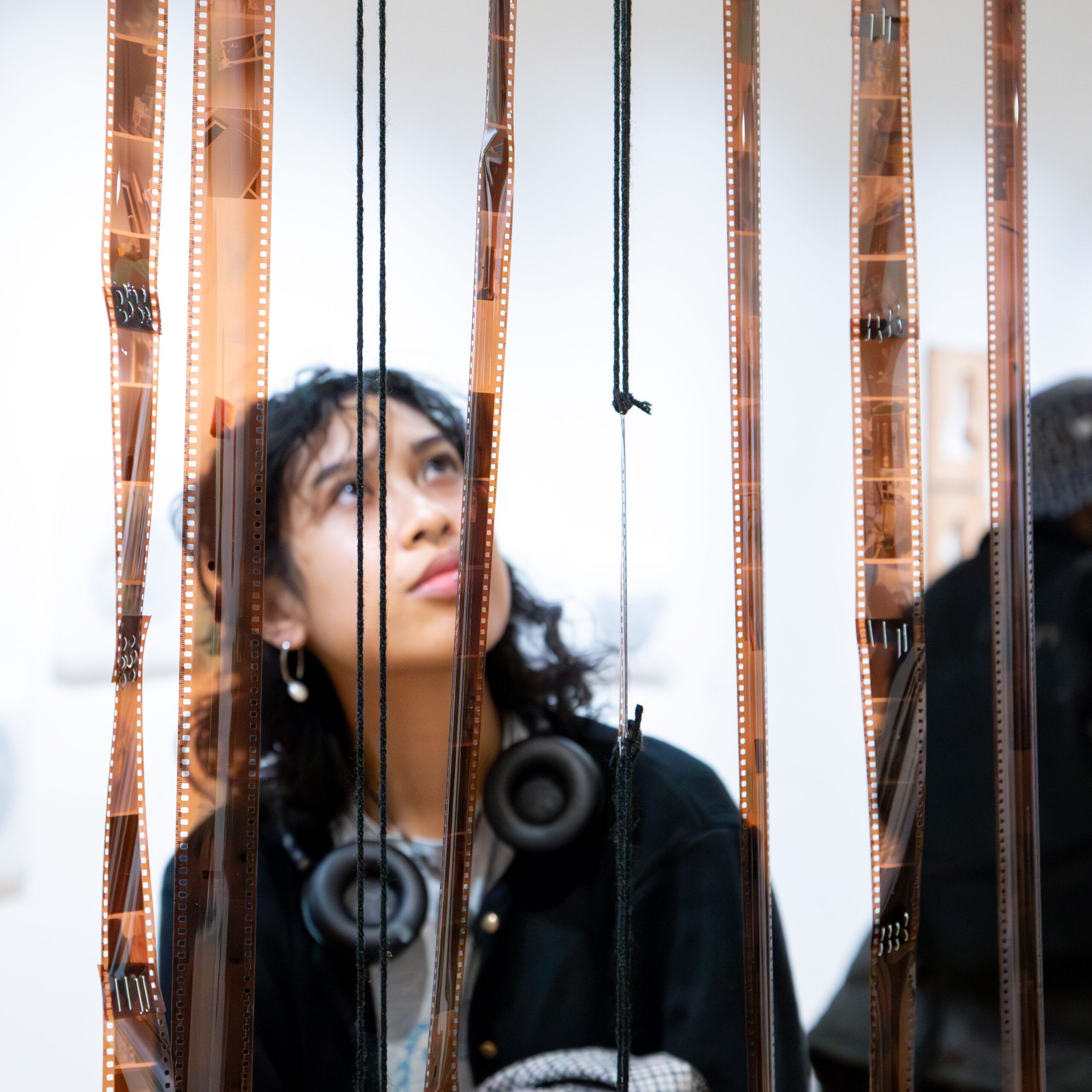 A student in a gallery space is looking at a fellow student's installation of film negatives hanging down from the ceiling like streamers.