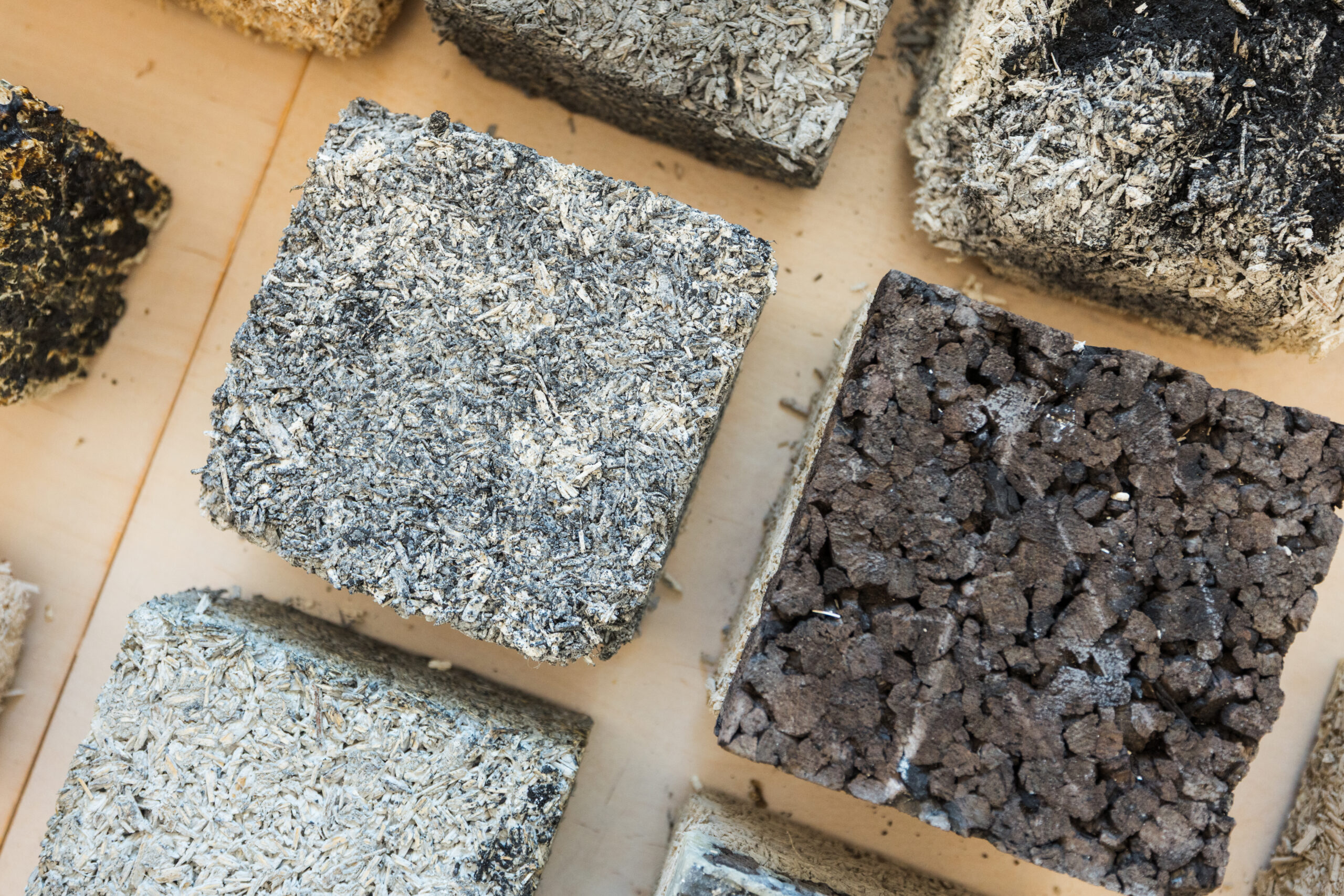 Close-up photo of nine textured building material samples arranged in a grid on a light wooden surface. The blocks are made from various sustainable and decarbonized mixtures, including fibrous hempcrete, compressed earth, and recycled aggregates. The materials have varying colors and compositions—ranging from tan and straw-like to dark and porous—demonstrating experimental approaches to eco-friendly construction techniques.