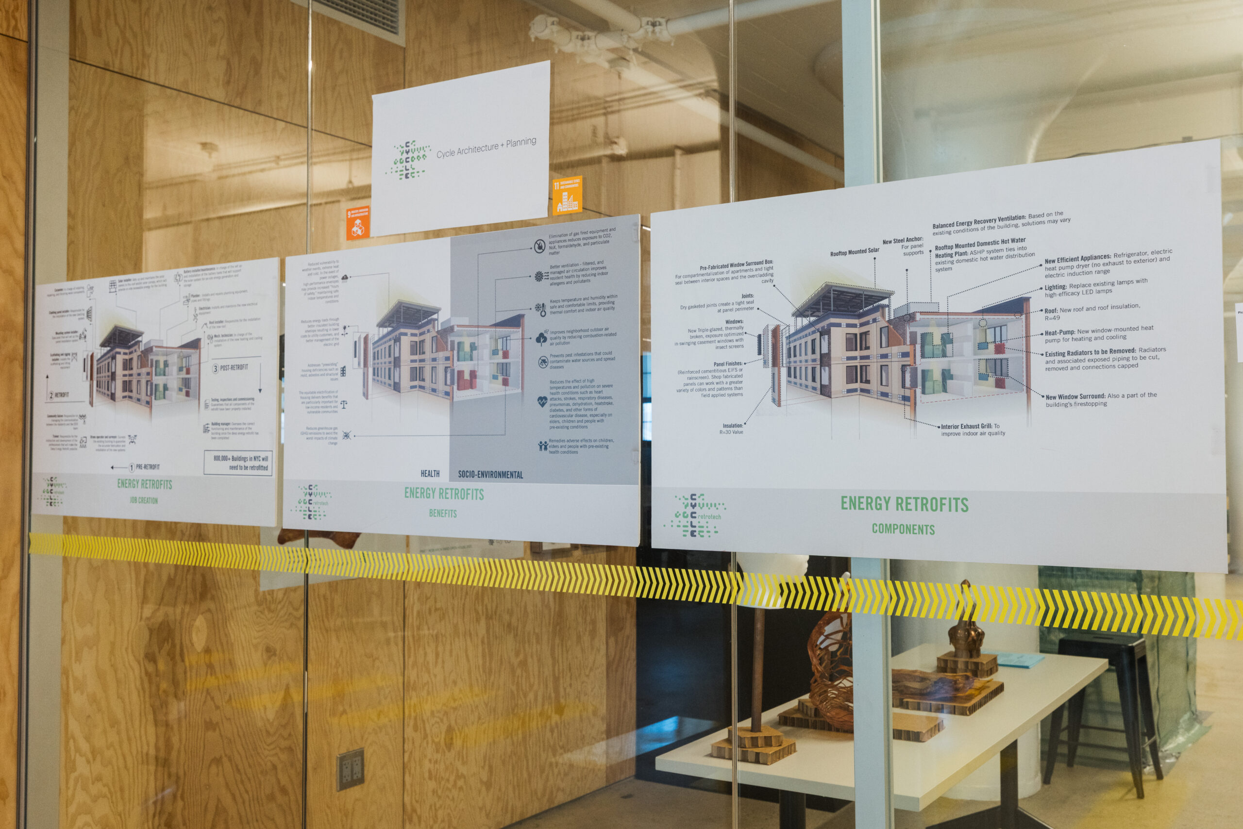 Photo of a glass wall exhibit featuring a presentation on energy retrofits by Cycle Architecture + Planning. Three large posters display annotated diagrams of a multi-unit residential building with labeled retrofit features such as new insulation, solar panels, efficient appliances, and ventilation systems. Text highlights health, environmental, and energy-saving benefits. A yellow safety stripe runs across the glass, and a table inside holds wooden architectural models and pamphlets.