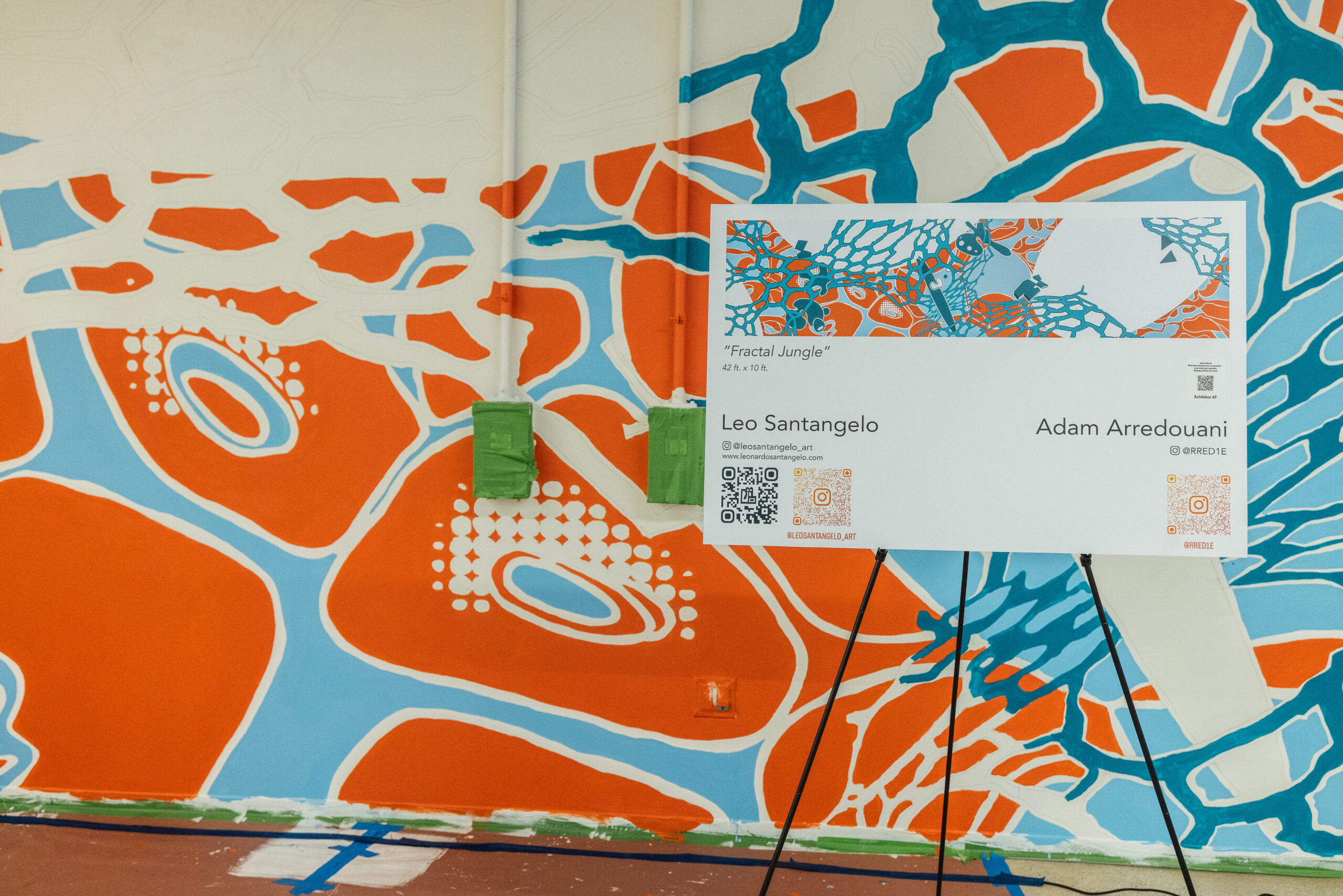 Close-up view of the nearly finished “Fractal Jungle” mural, showcasing dynamic biomorphic forms in orange, blue, and white. A printed sign in front of the wall provides information about the piece, listing its dimensions as 42 x 10 ft and crediting artists Leo Santangelo and Adam Arredouani. QR codes and social media handles are included at the bottom of the sign, which mirrors the mural's color scheme and graphic style.