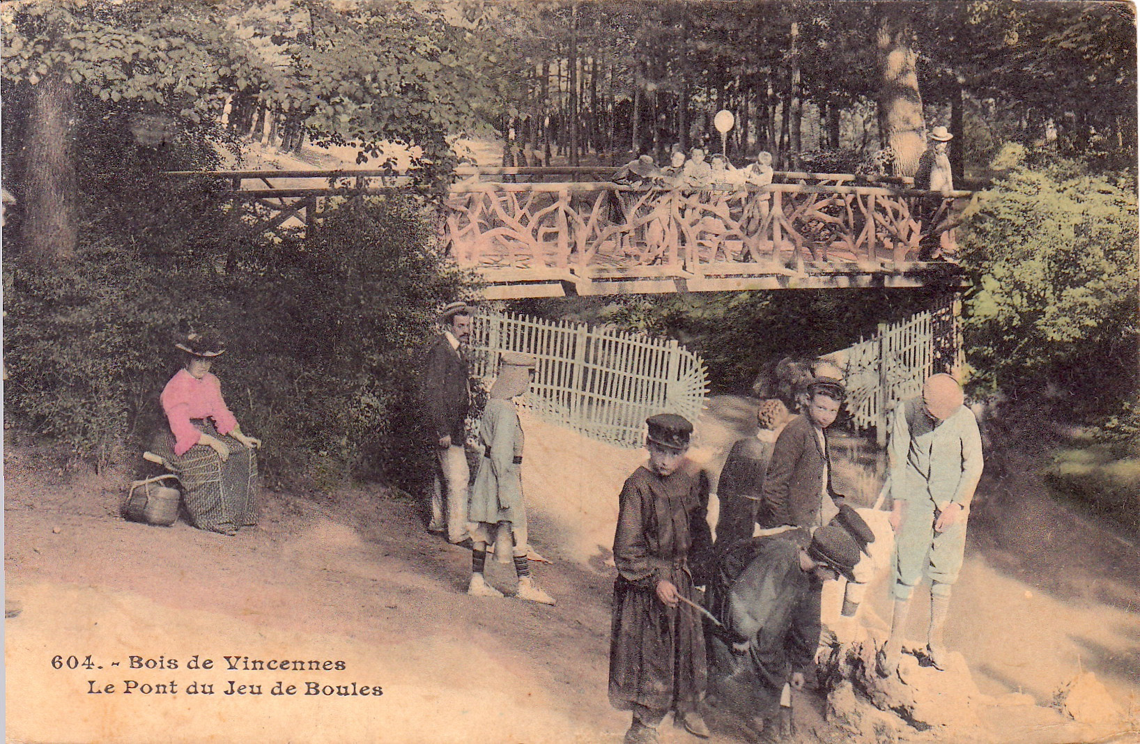 Hand-tinted vintage postcard showing a wooded park scene in Bois de Vincennes, France. The setting includes a rustic bridge with people leaning on the railing and a group of children and adults playing or walking below. A seated woman in a pink blouse observes from the foreground.