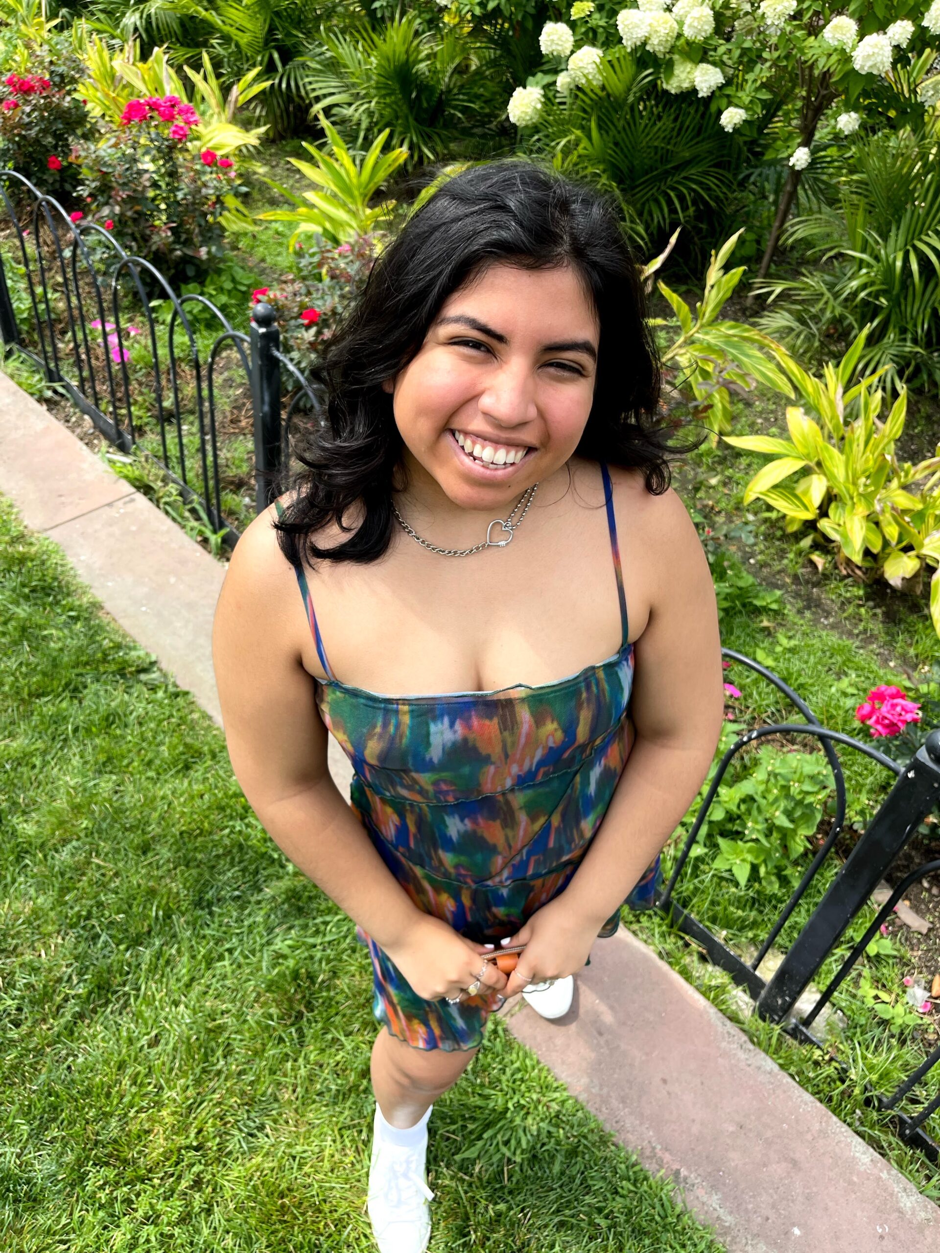 A smiling young woman in a colorful dress standing on a grassy path with blooming flowers and lush greenery in the background.