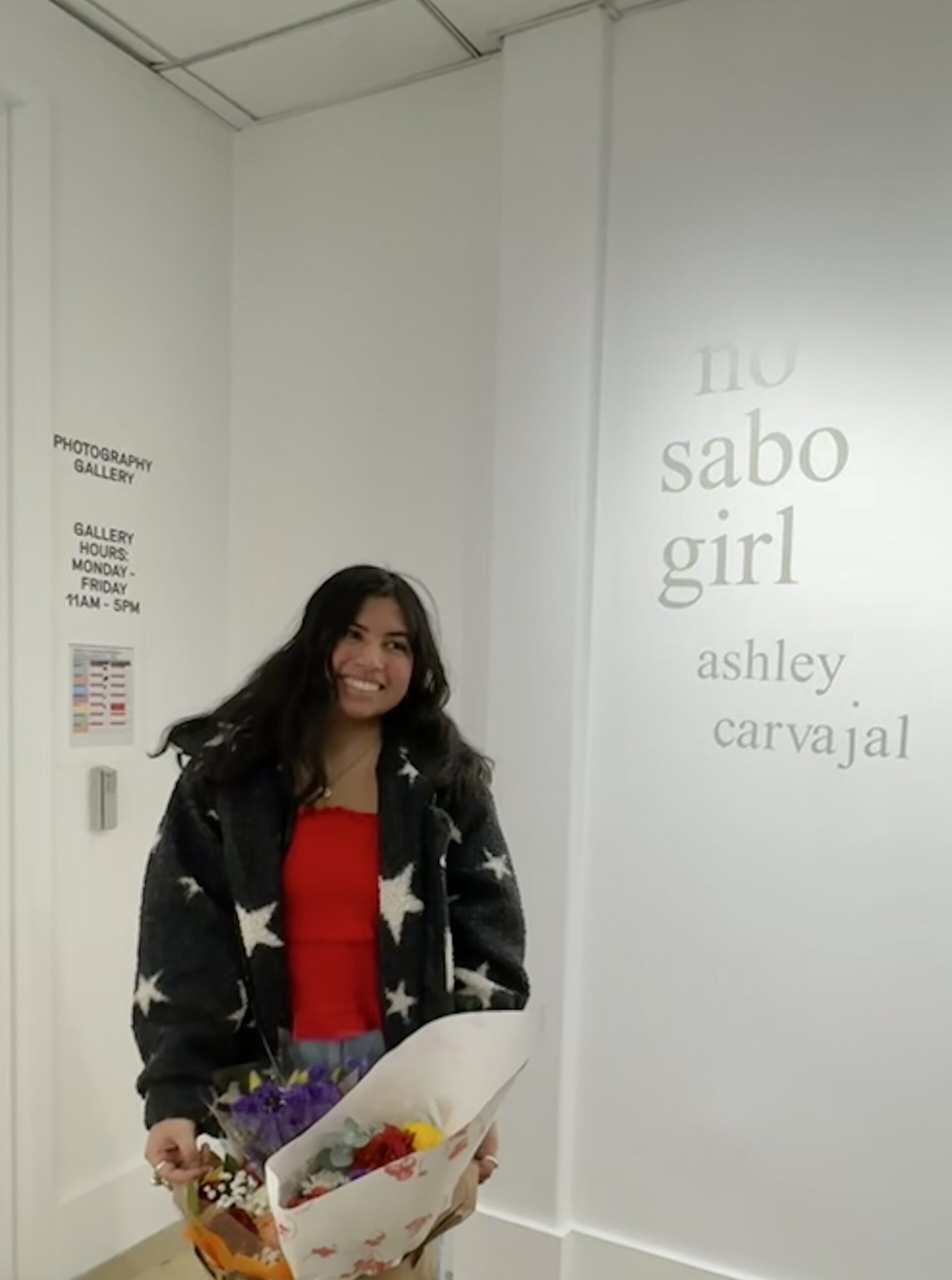 A woman holding a bouquet of flowers standing in front of a white wall displaying the exhibition title “no sabo girl” and the name “ashley carvajal.”