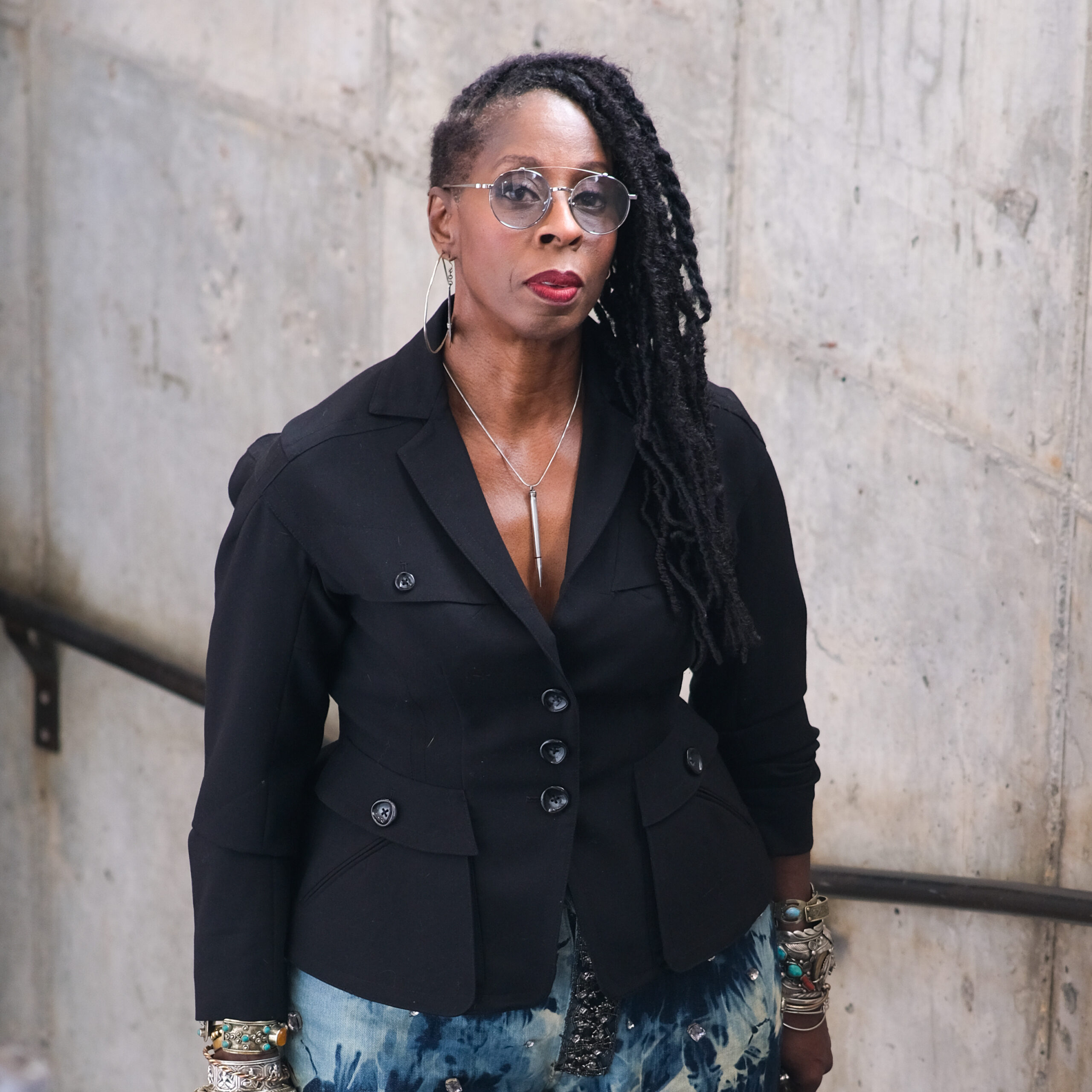 Portrait photo of a woman with dark skin, long dreadlocks swept to one side, and wearing round glasses. She is dressed in a fitted black blazer with multiple buttons and patterned blue-and-white pants. She accessorizes with layered silver bracelets, hoop earrings, and a long pendant necklace. The background is a concrete wall, suggesting an urban setting, and she looks confidently toward the camera. Photo credit: Anthony Artis.