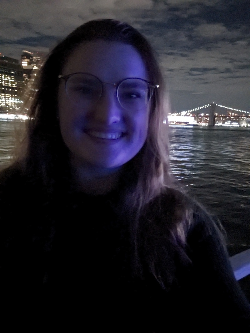 A woman smiling at night with purple lighting on her face, standing near the water with a lit-up bridge and city skyline in the background.