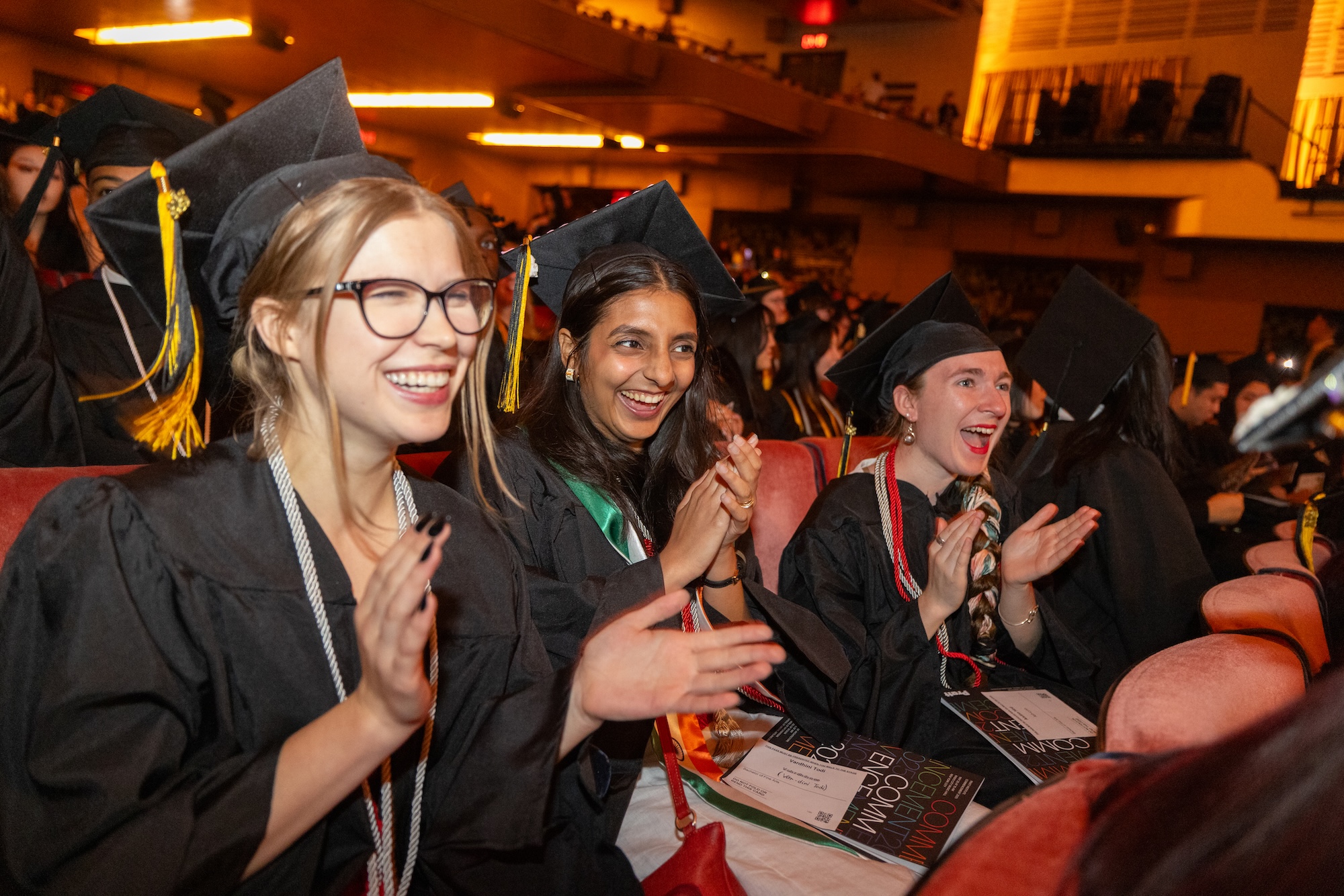 Graduates in caps and gowns sit in an auditorium, smiling and clapping enthusiastically during a commencement ceremony.