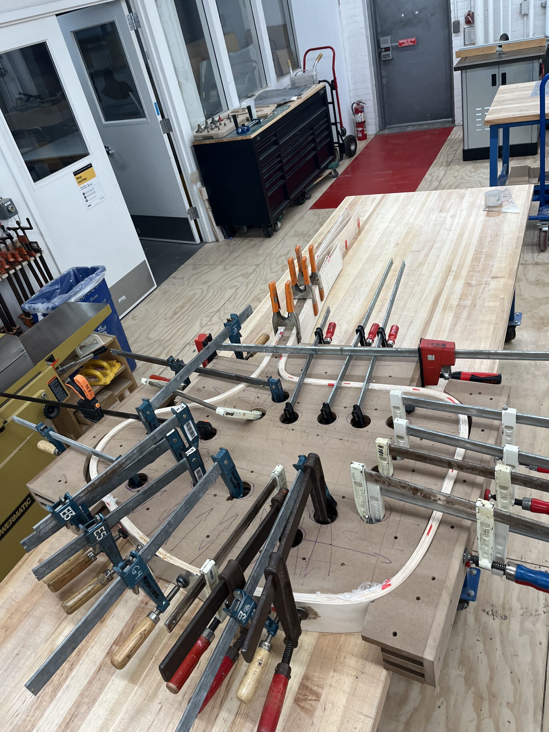 A woodshop workspace with multiple clamps securing curved laminated wood pieces to a large tabletop jig. The room includes various tools, stools, and benches, with a red safety mat near a closed metal door. The setting appears to be part of a university or professional fabrication lab.