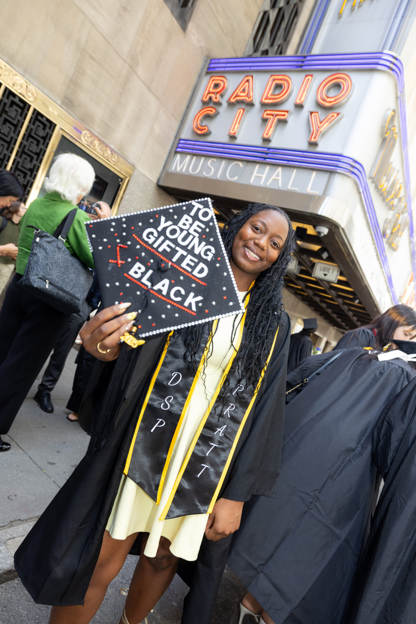 A graduate in cap and gown proudly displays a cap that reads “TO BE YOUNG, GIFTED, & BLACK” in rhinestones, standing outside Radio City Music Hall.