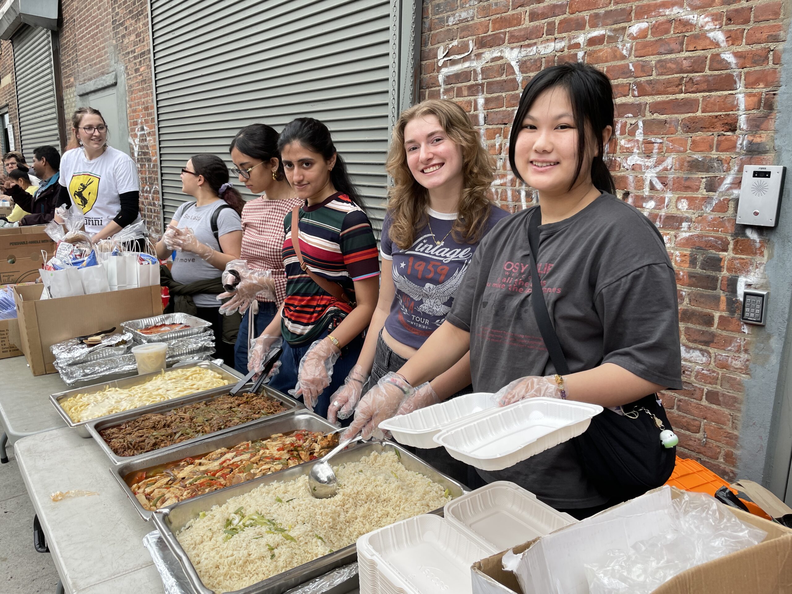 A group of young volunteers stands behind a table lined with large metal trays of hot food, ready to serve meals. The dishes include pasta with Alfredo sauce, stir-fried vegetables, a chicken and pepper dish, and a tray of seasoned rice. One volunteer in a dark t-shirt holds biodegradable food containers, smiling at the camera. Next to her, others are wearing plastic gloves, holding serving utensils, and preparing to dish out food. In the background, two volunteers manage a table of care packages and snacks. The scene is set outdoors against a brick wall with graffiti, with a metal roll-up door to the side.