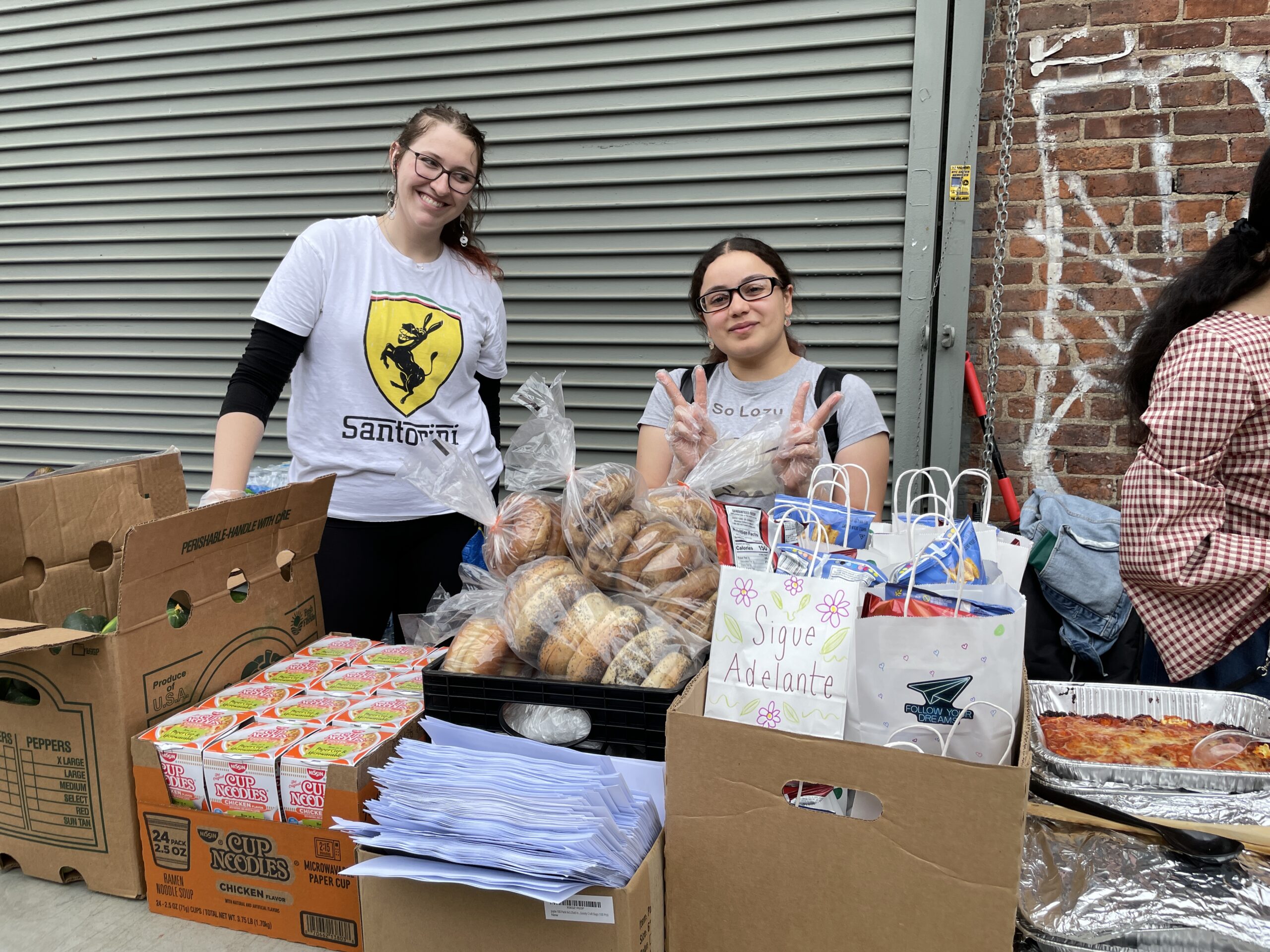 Two young women stand behind a table filled with food and care packages, smiling at the camera. The woman on the left wears a white t-shirt with a yellow “Santorini” logo and glasses, while the woman on the right holds up both hands in peace signs, wearing clear plastic gloves and a grey t-shirt that reads "So Lazy." The table is loaded with boxes of Cup Noodles, crates of bagels in plastic bags, a stack of printed flyers, and several decorated white gift bags labeled with phrases like "Sigue Adelante" (Spanish for "Keep Going"). To the right, aluminum trays of prepared hot food are visible. The scene is set outdoors, against a metal roll-up door and a brick wall with graffiti.
