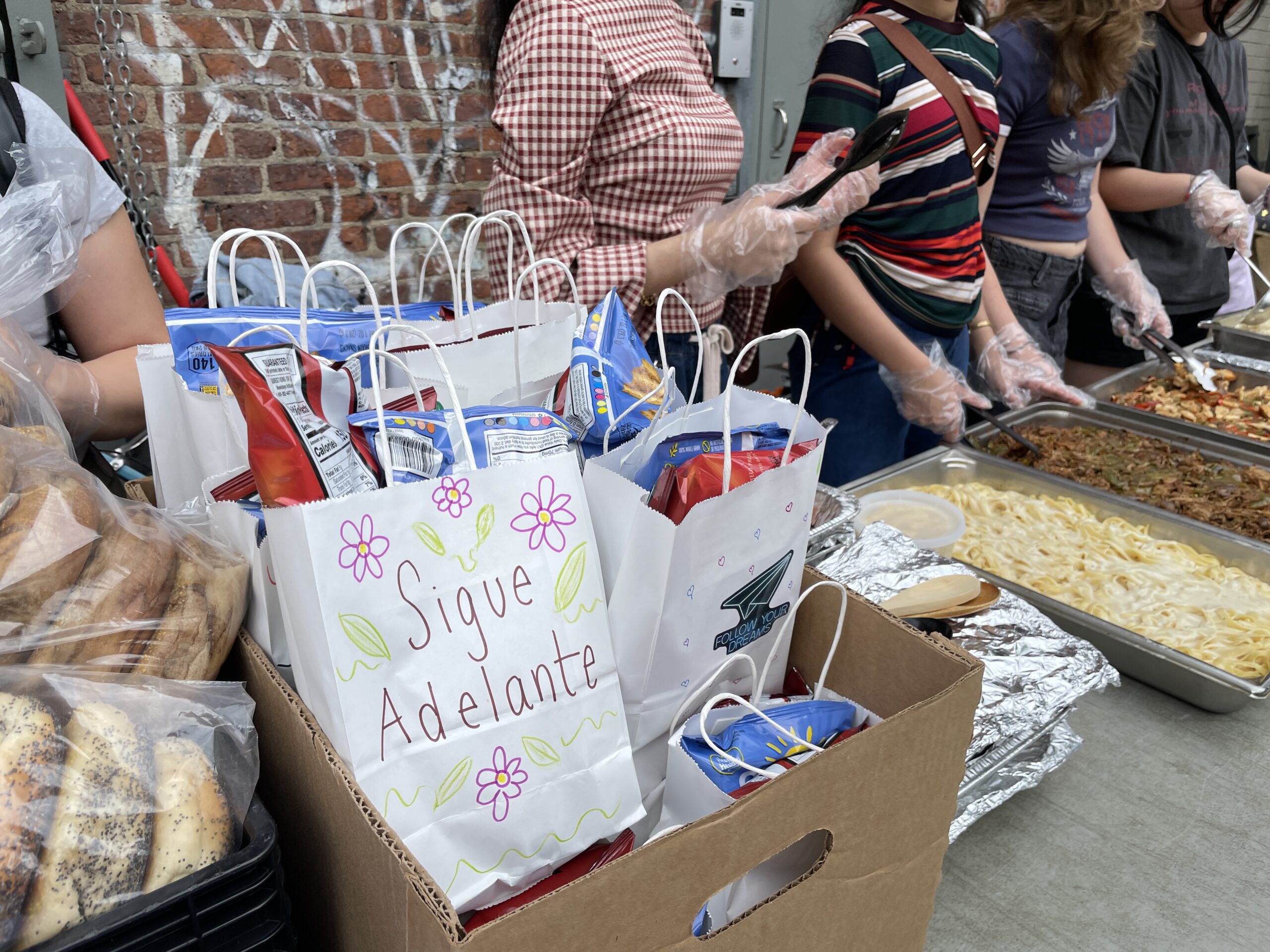A row of volunteers wearing plastic gloves is serving hot food from large metal trays at an outdoor event. The dishes include pasta with Alfredo sauce and a mixed vegetable stir-fry. In the foreground, there are cardboard boxes filled with decorated white paper bags containing assorted snacks, including bags of chips. One bag prominently displays the message "Sigue Adelante" (Spanish for "Keep Going") with hand-drawn flowers and leaves. To the left, there are plastic bags filled with bagels stacked in a crate. The background shows a brick wall with graffiti.