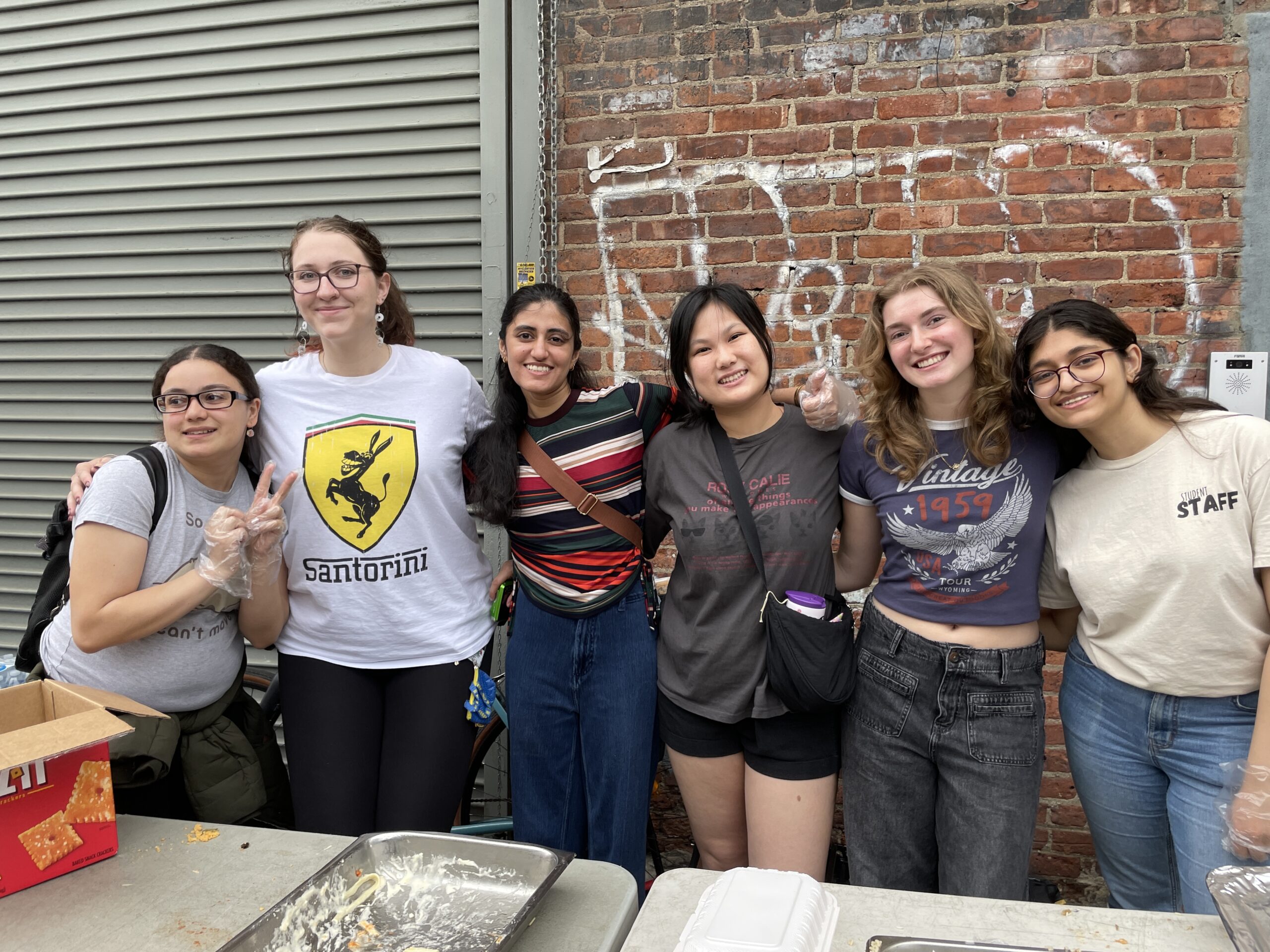 A group of six young women stands smiling in front of a brick wall with graffiti, posing together behind a table of food trays at an outdoor volunteer event. They have their arms around one another and appear cheerful and relaxed. Some wear food service gloves, indicating participation in food distribution. From left to right: the first person wears a gray T-shirt and glasses, the second wears a white “Santorini” shirt with a donkey logo, the third wears a striped top with blue jeans, the fourth wears a dark gray T-shirt and crossbody bag, the fifth wears a navy blue “Vintage 1959” shirt and black jeans, and the sixth wears a “Staff” T-shirt and glasses. Behind them is a corrugated metal door and a red brick wall.