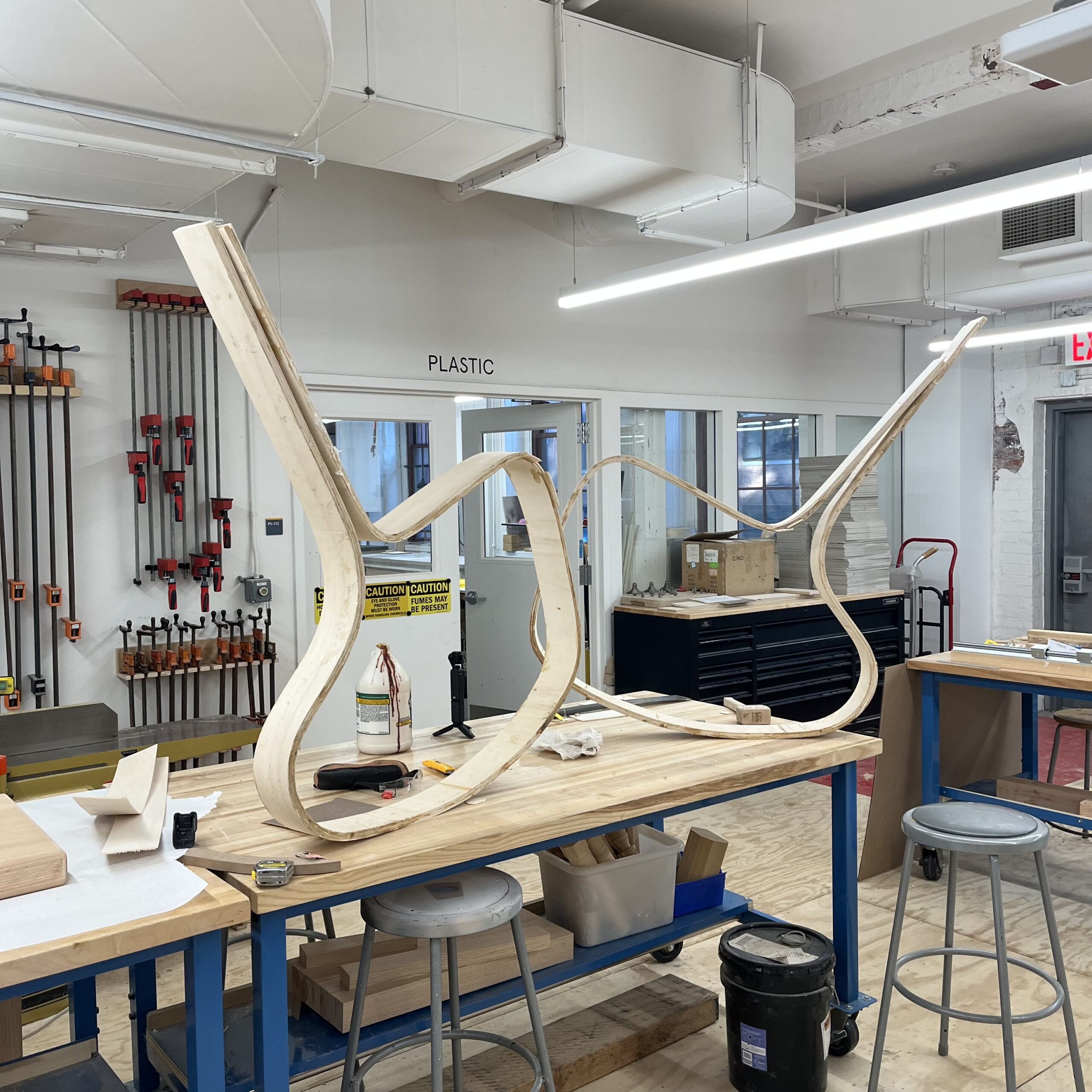 Two large, curved plywood frame components resting upside down on a workbench in a fabrication studio. The bent wood structures are in progress, suggesting a furniture-building process involving lamination and steam bending. Tools and clamps hang on the wall in the background.