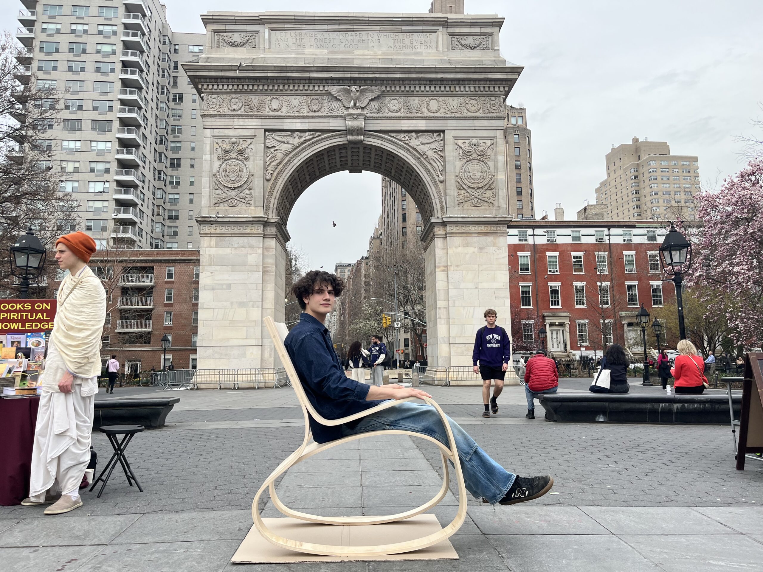 A young man seated in a modern plywood rocking chair outdoors at Washington Square Park in New York City. The iconic marble archway is visible behind him, surrounded by pedestrians and blooming trees. A vendor table with books on spiritual knowledge is positioned nearby.