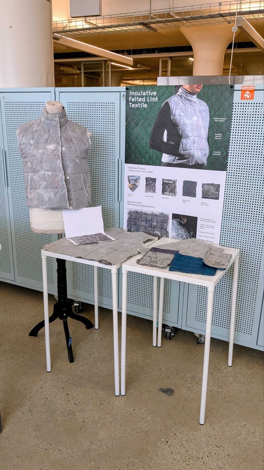A display titled “Insulative Felted Lint Textile” features a mannequin wearing a quilted gray vest made from repurposed dryer lint. Beside it, two tables showcase textile samples in various textures and colors, along with a large poster explaining the fabrication process and design concept.