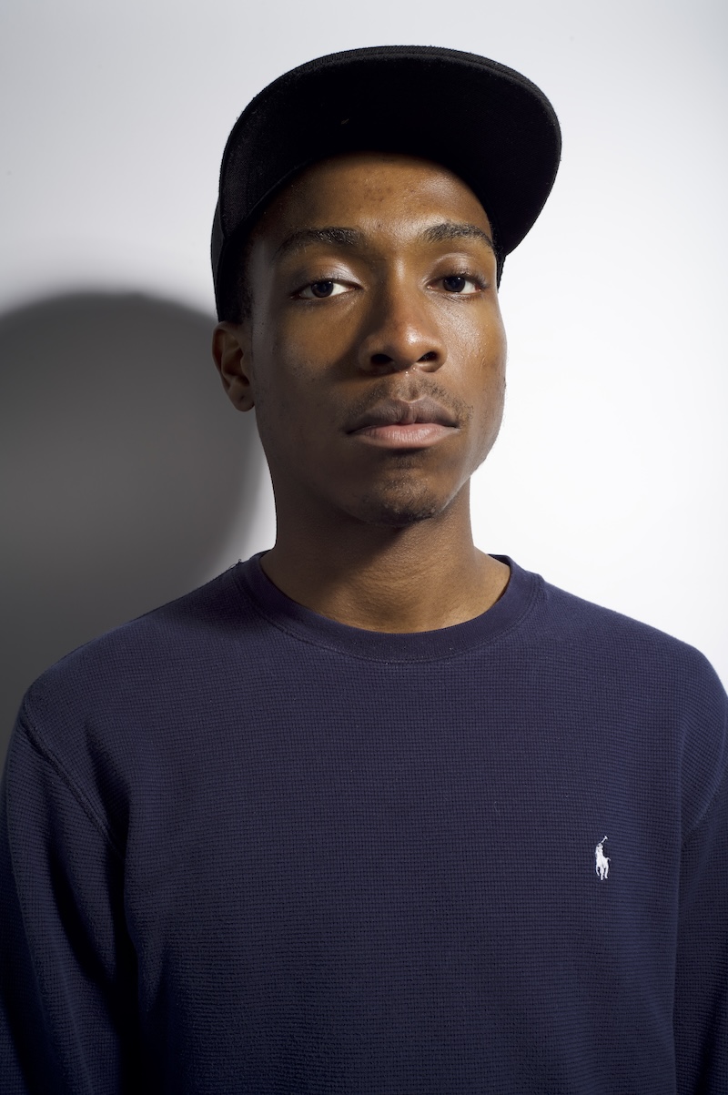 A formal portrait of a man in a dark navy shirt and black cap, looking at the camera against a light background.