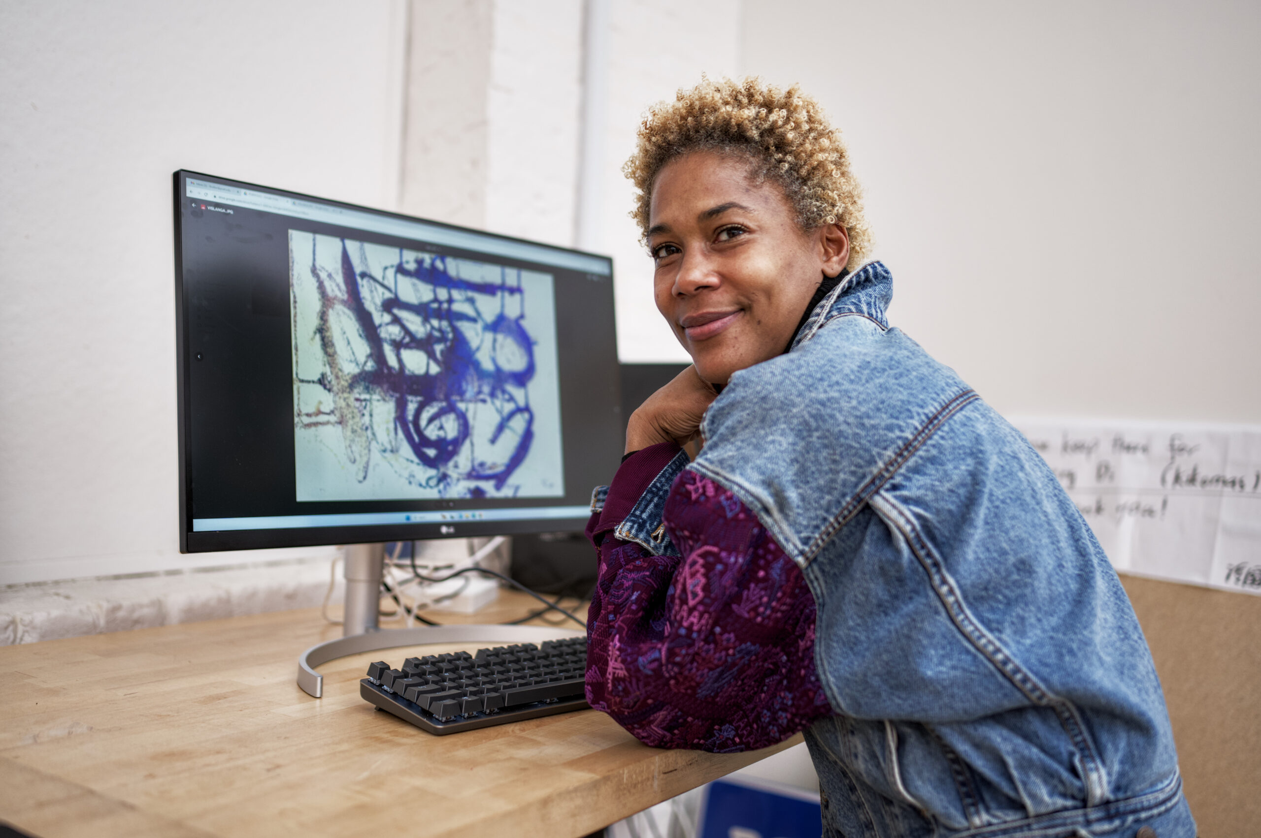 Student sitting at a desk and smiling while showing digital artwork on a computer screen
