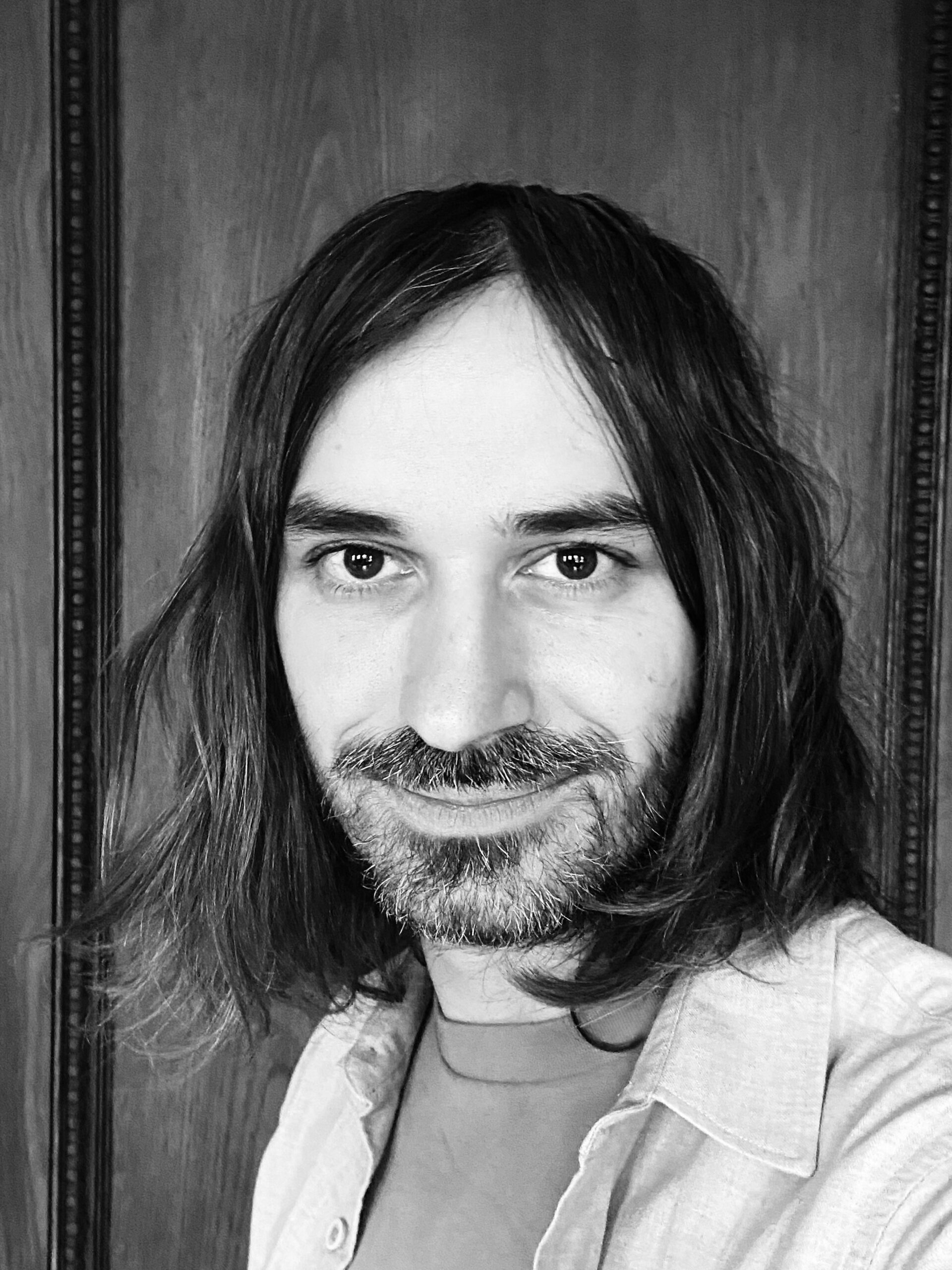 A black and white headshot of a man with shoulder-length hair looking at the camera.