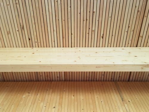 A close-up view of a wall and bench constructed from nail-laminated timber (NLT), featuring vertically aligned, narrow wooden slats with a light, natural finish. The clean, uniform texture emphasizes the modular aesthetic and precision typical of mass timber construction.