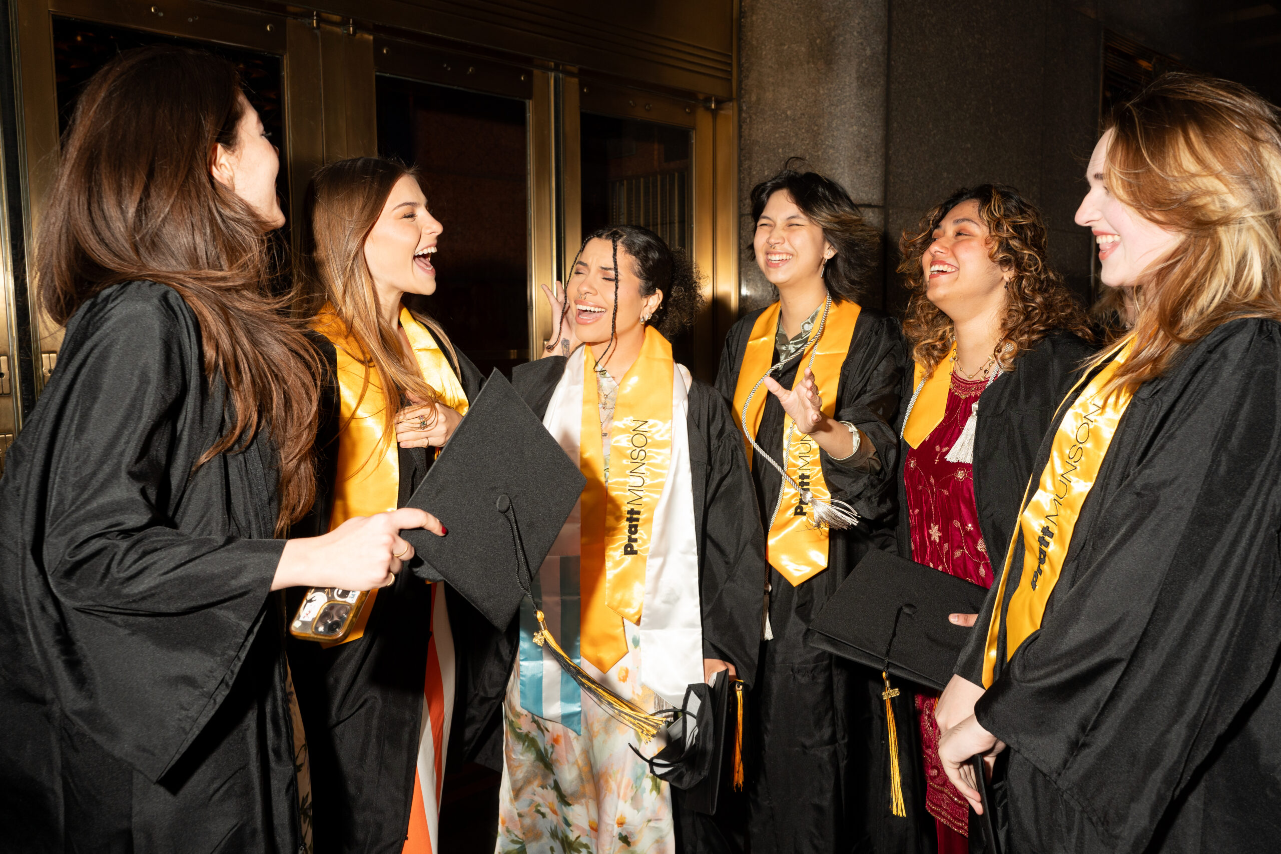A group of six graduates wearing black gowns and gold "Pratt Munson" sashes stand outside, laughing and enjoying the moment together.