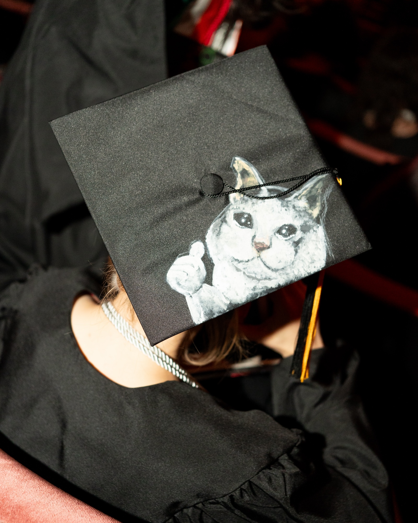 A graduation cap decorated with a hand-painted white cat giving a thumbs-up gesture.