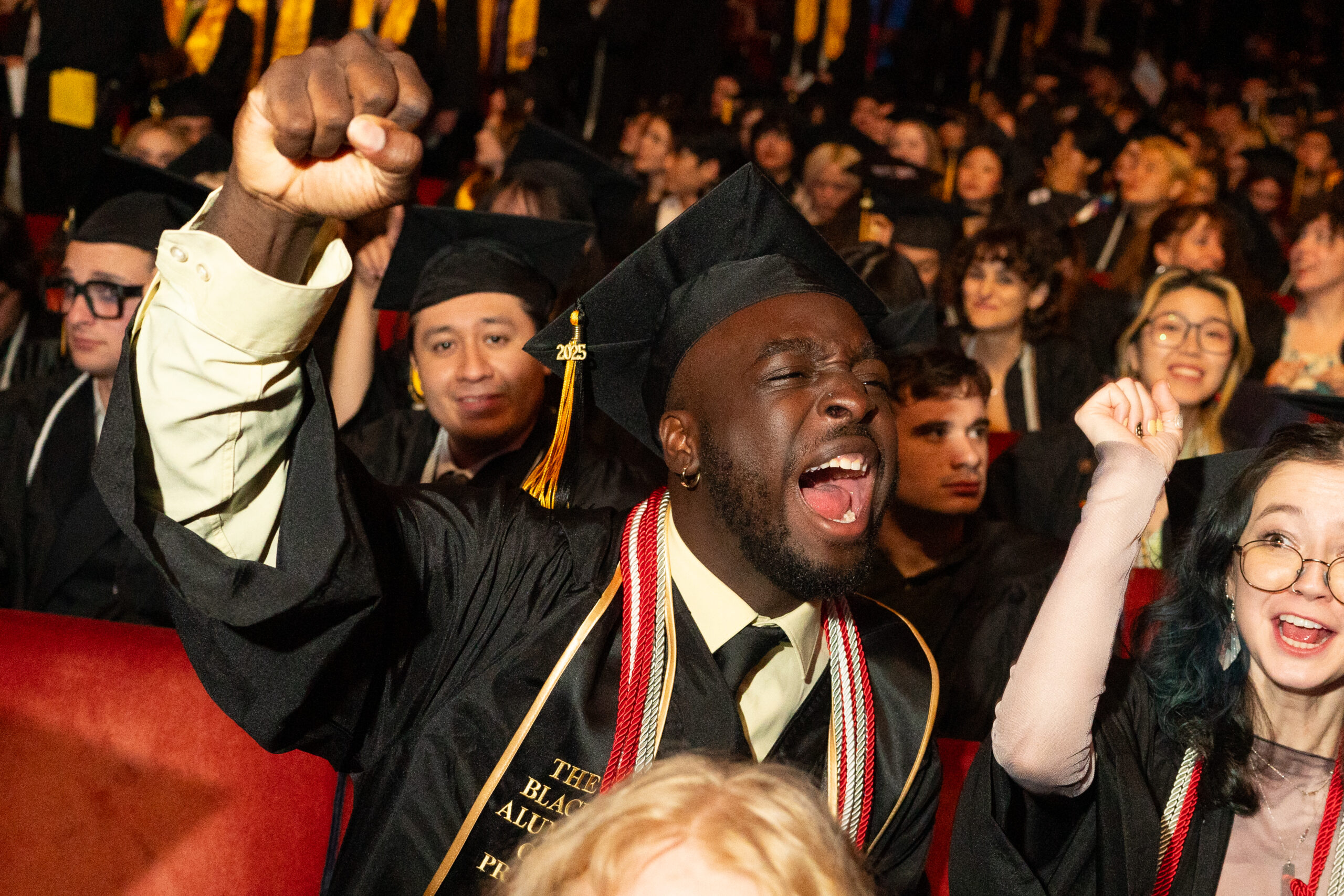 A joyous graduate in a cap and gown raises a fist in celebration while seated among fellow students at a commencement ceremony.