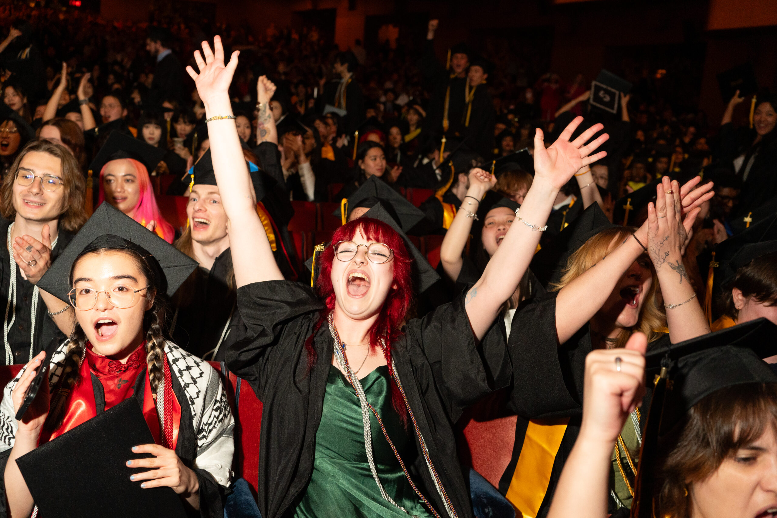 A group of excited graduates in caps and gowns clap and smile while seated in the auditorium.