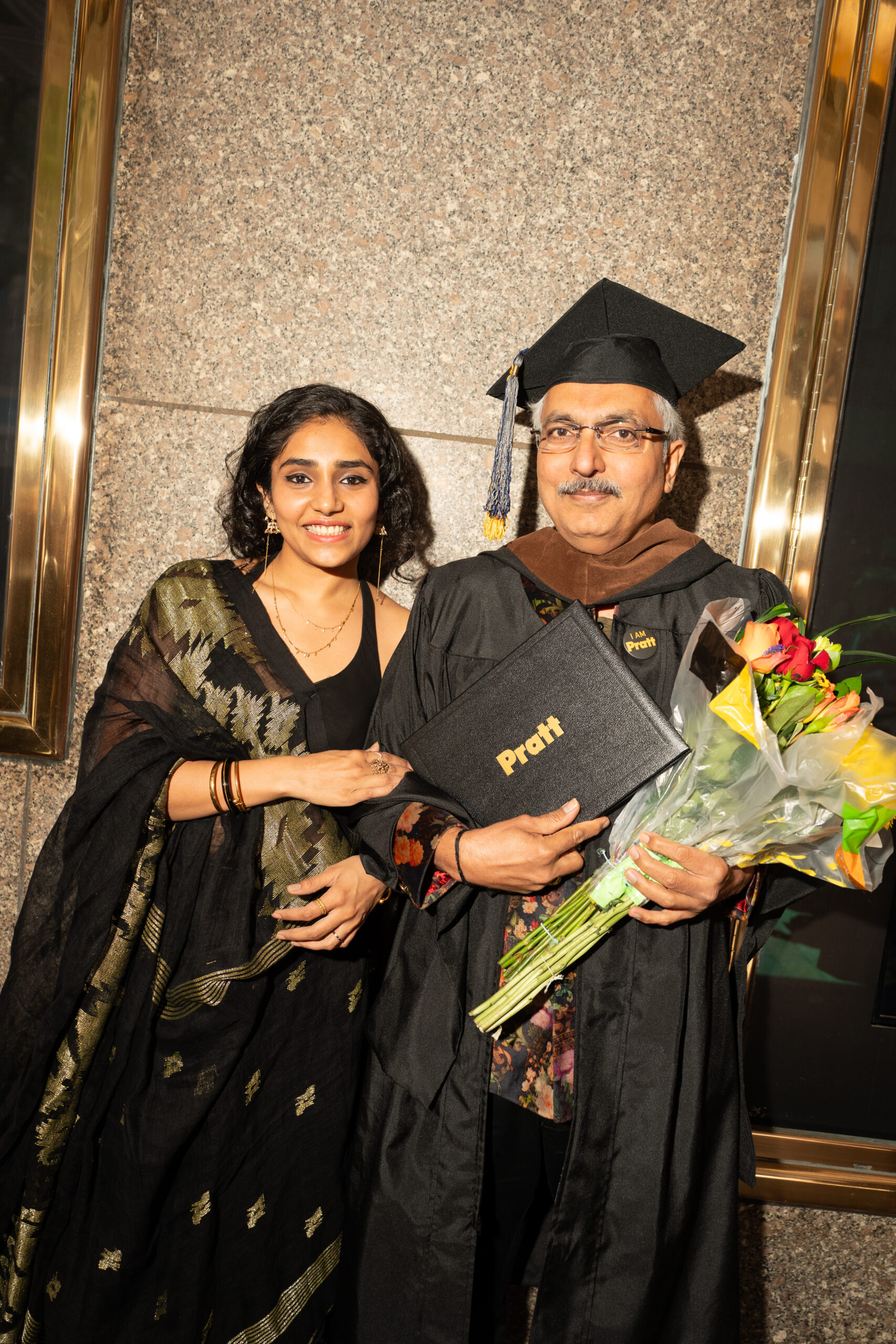 A graduate in a cap and gown holds a diploma and bouquet while standing with a smiling family member or friend.