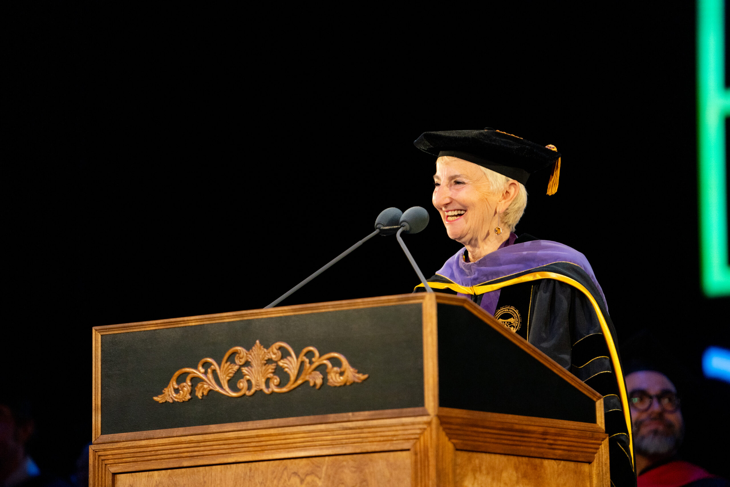 A smiling speaker in academic regalia delivers a speech at a graduation ceremony from behind a wooden podium with gold detailing.